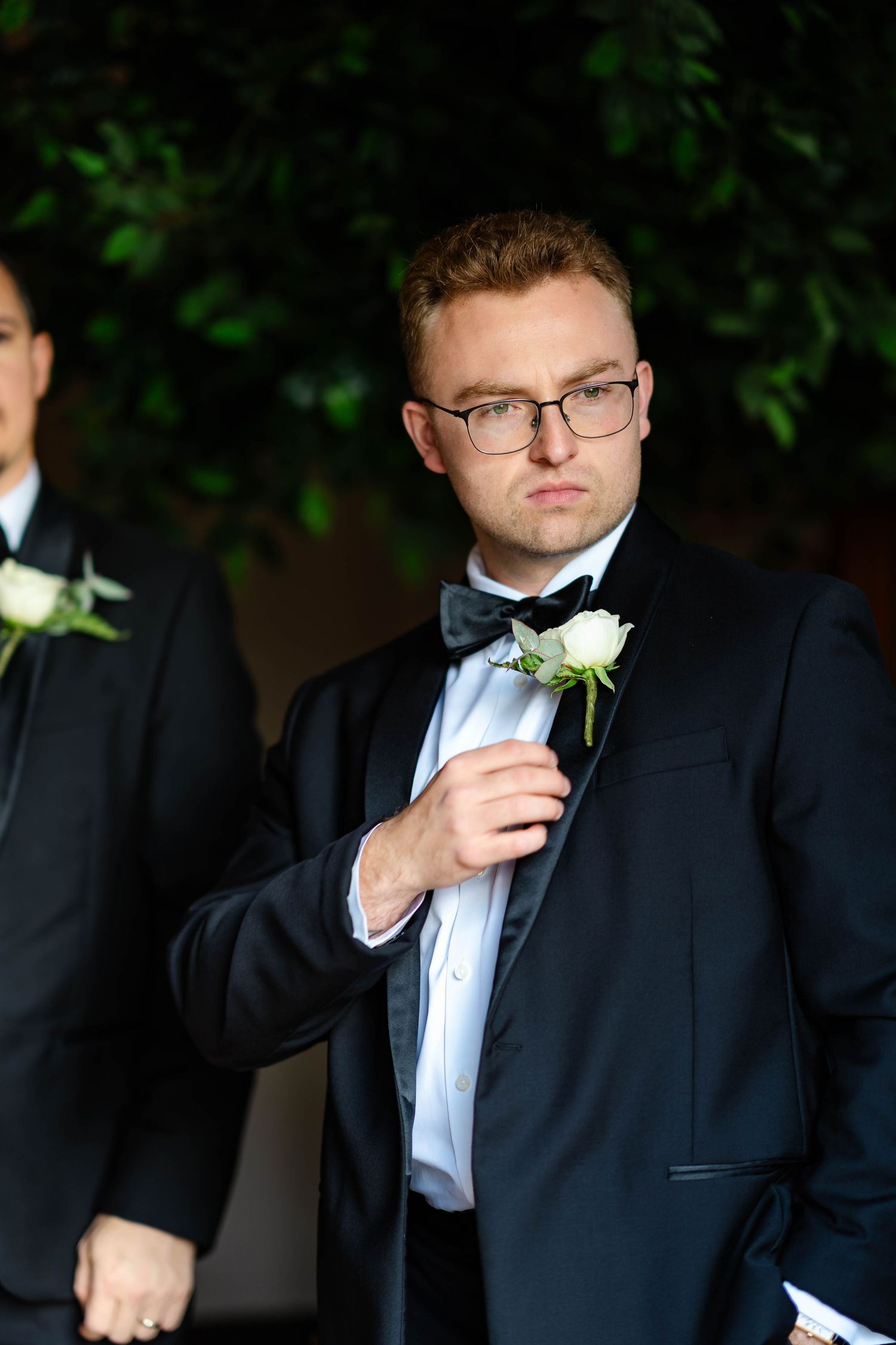 A groomsman in a black tuxedo adjusts his white boutonniere against a dark background.