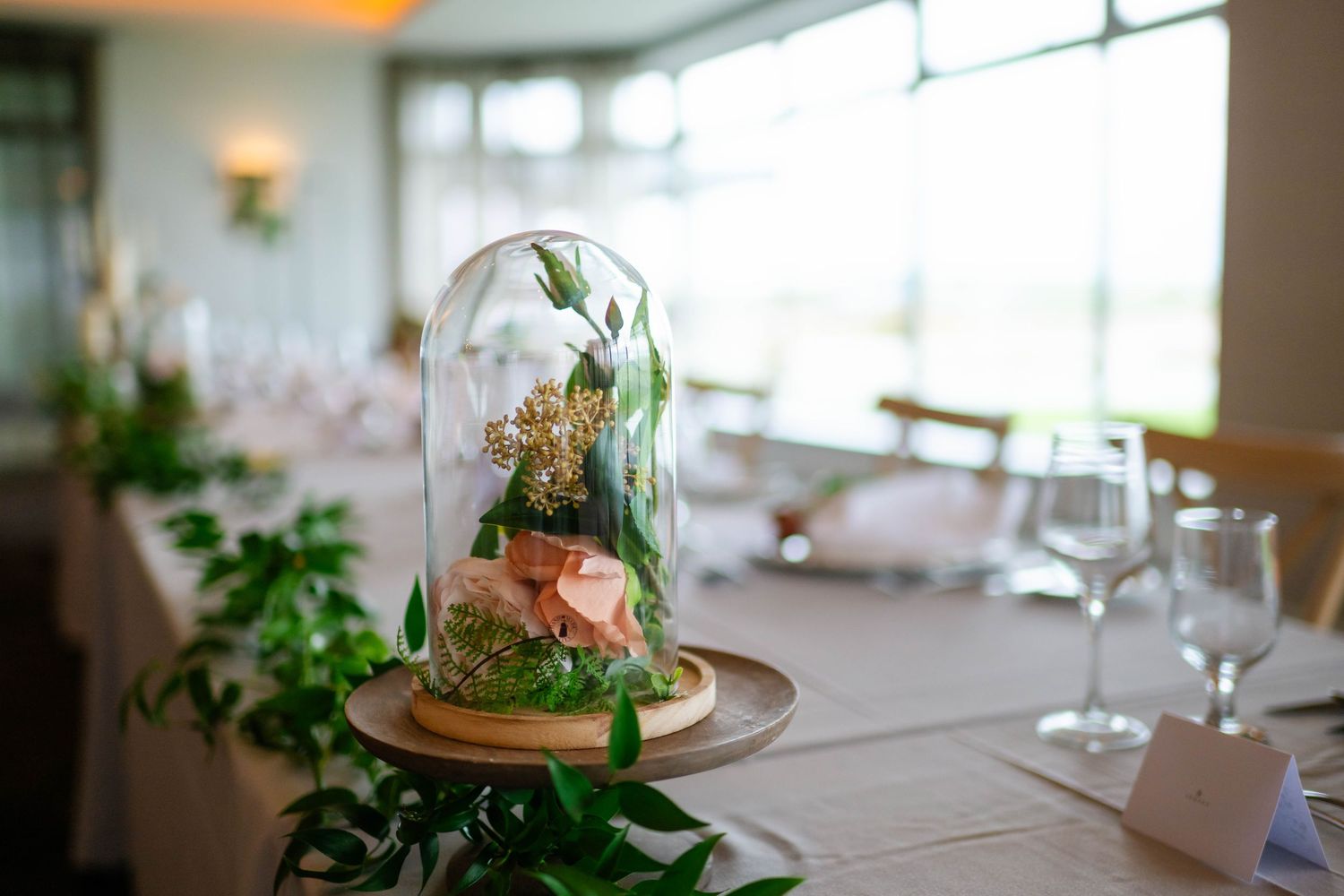 Elegant terrarium centerpiece with greenery under glass dome on wedding reception table.