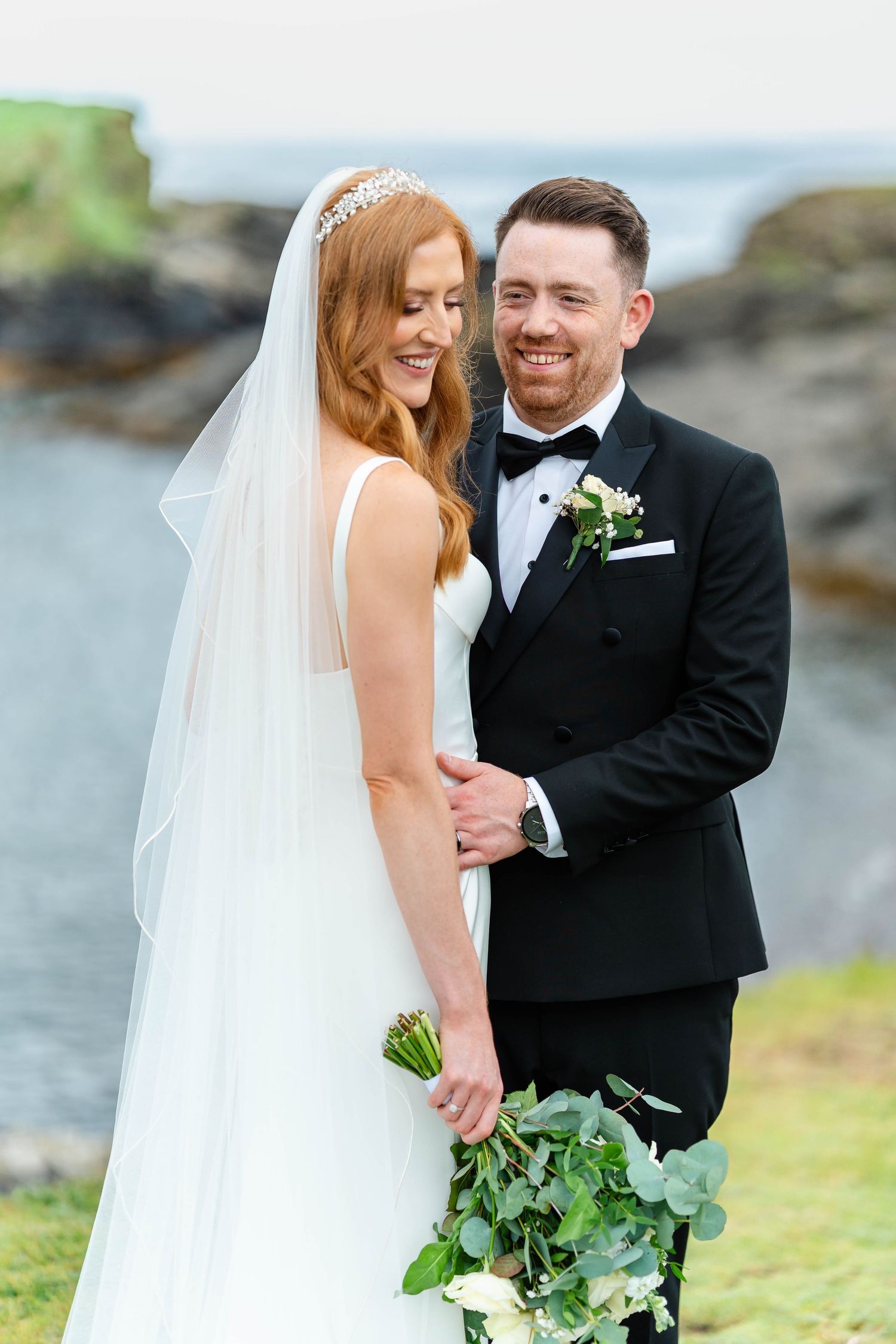 A romantic wedding moment captured against a rocky coastal backdrop with flowing white dress and formal attire.