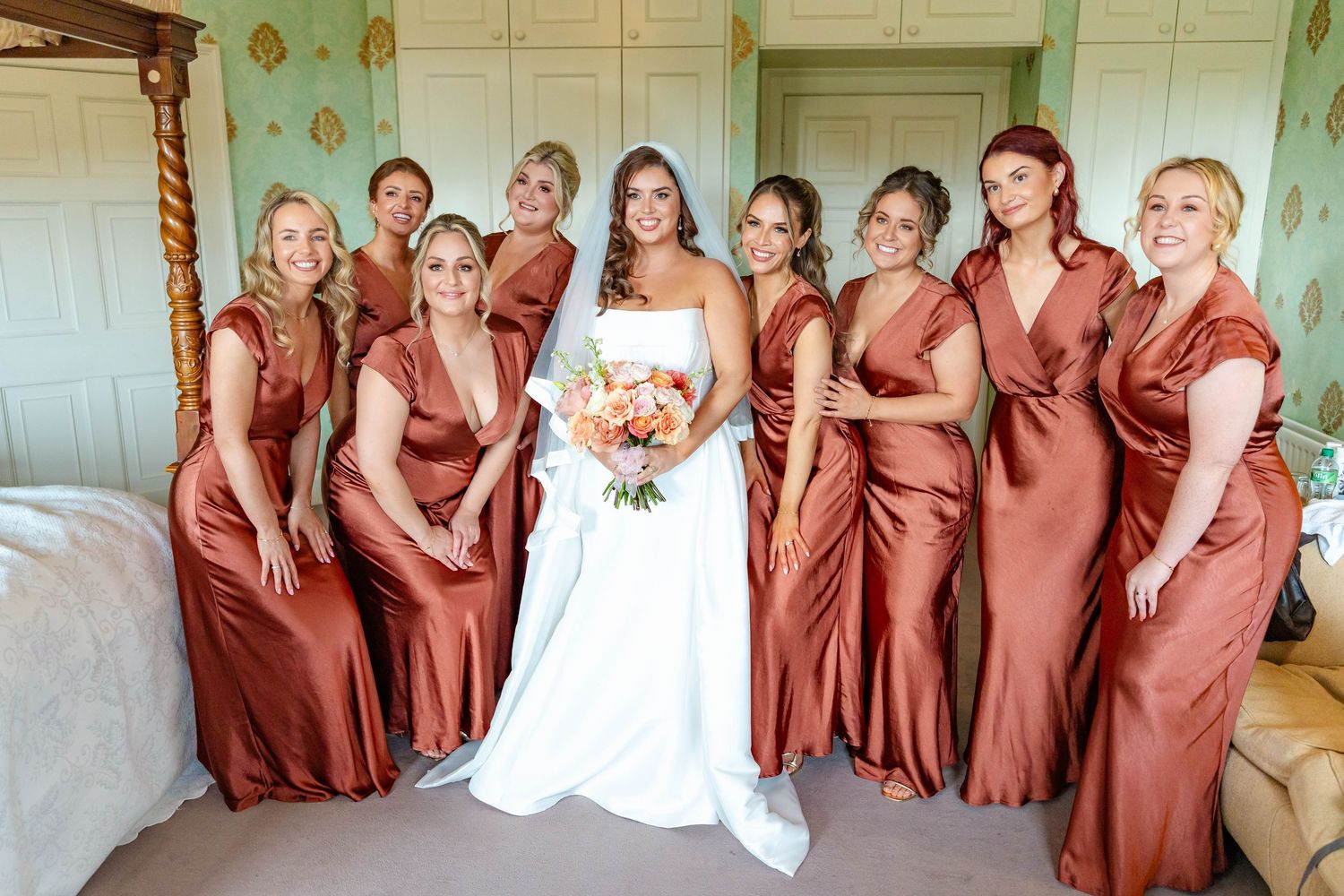 Bridesmaids in matching rust-colored satin dresses pose with bride in white gown during wedding celebration.