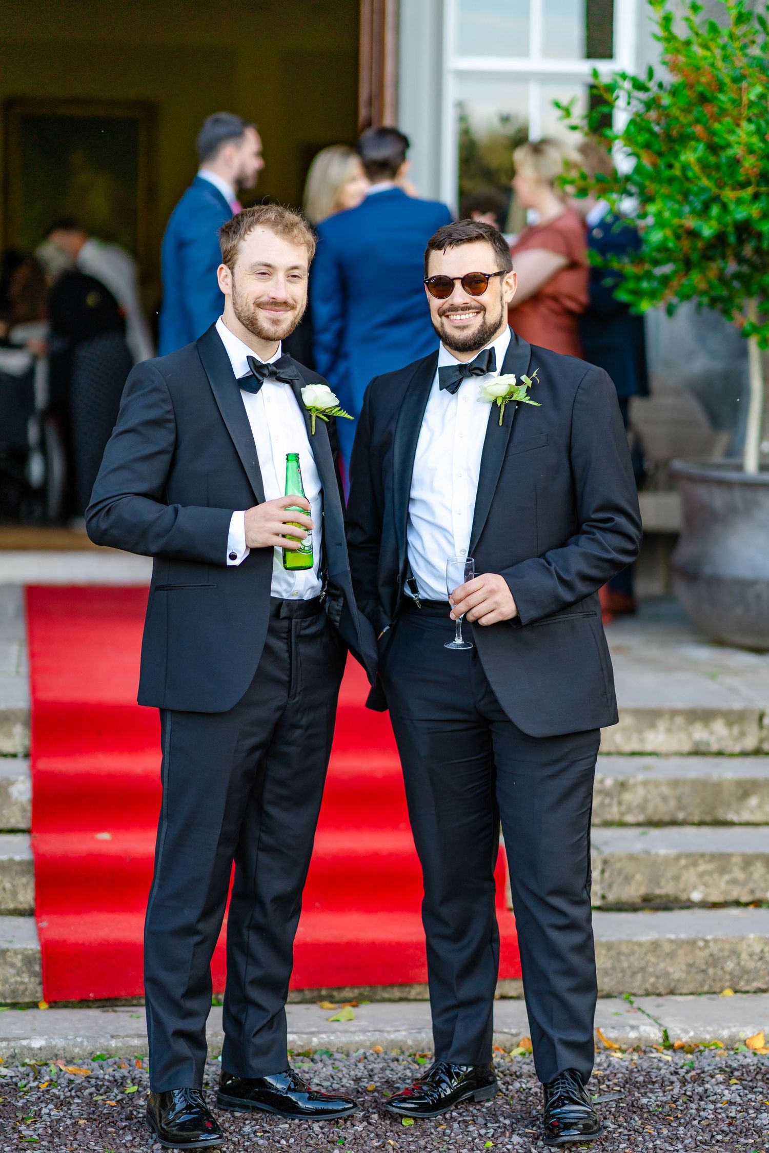 Two groomsmen in black tuxedos and bowties share a laugh while holding drinks on a red carpet at a wedding.