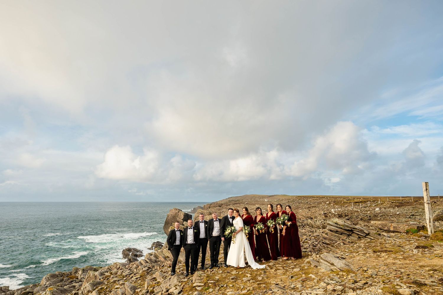 Panoramic view of wedding party on rugged coastal cliffs under cloudy skies.