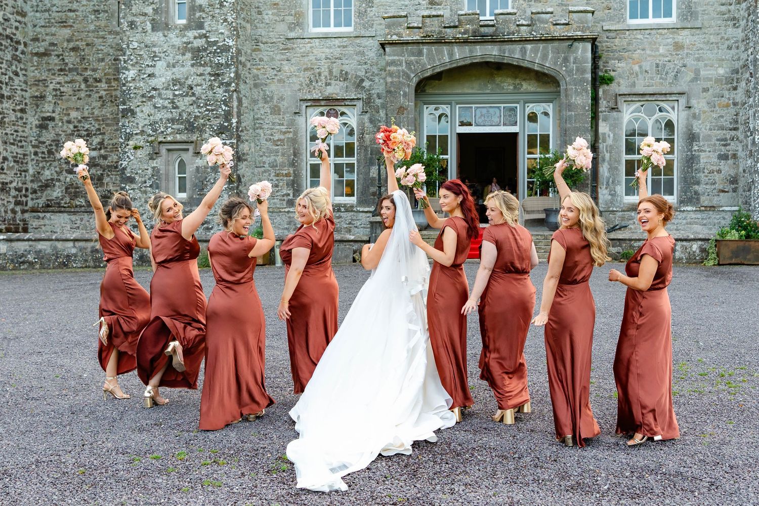Wedding party in rust-colored dresses poses playfully in sequence of celebratory jumping photos outside stone building.
