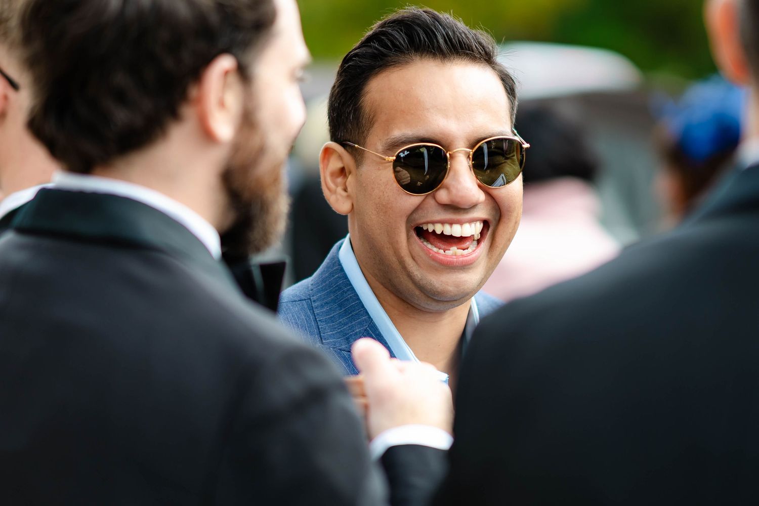 A group of people in formal attire laugh and smile at an outdoor social gathering.