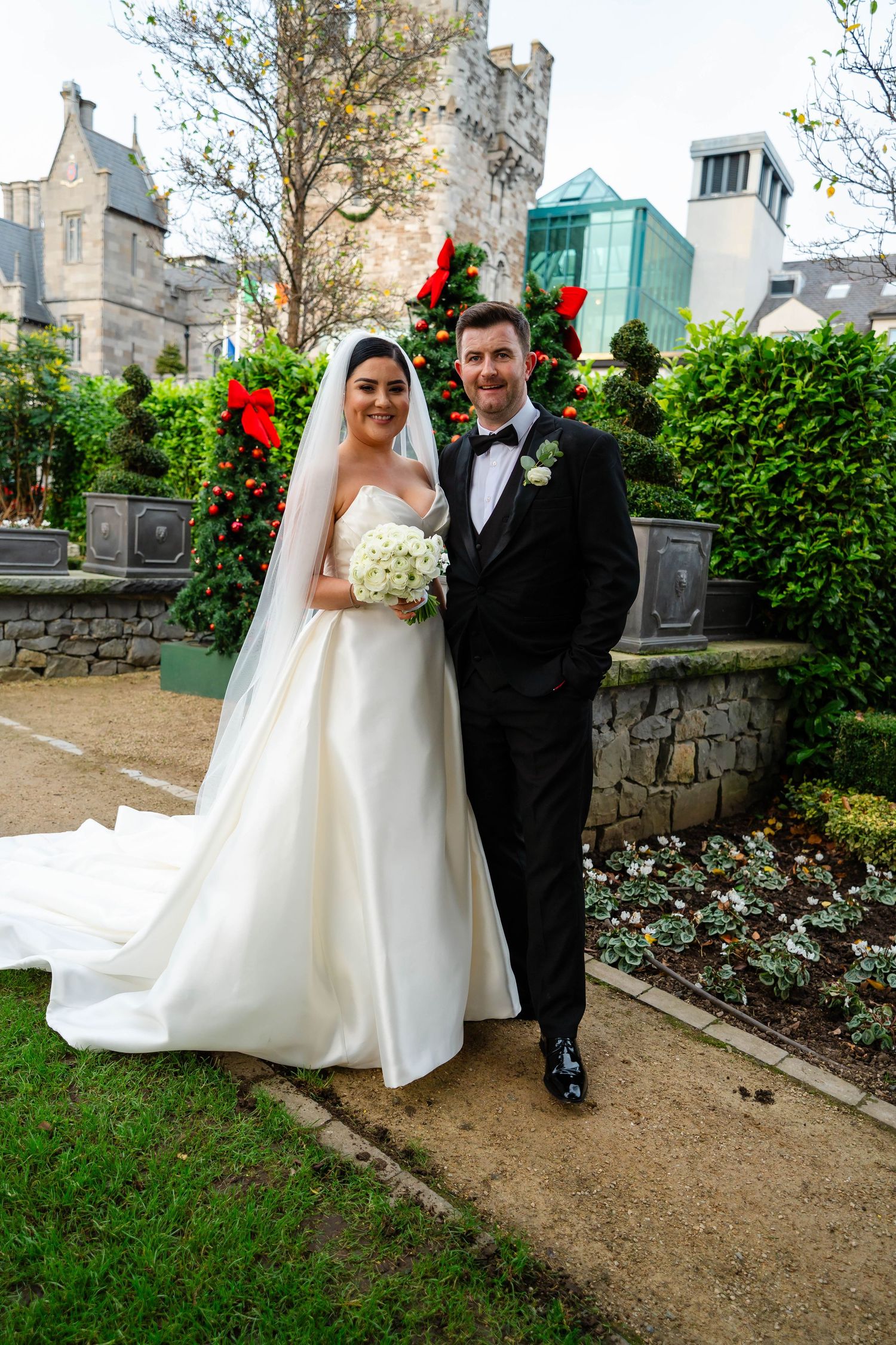 Newlyweds pose in garden with stone castle tower and greenery in background.