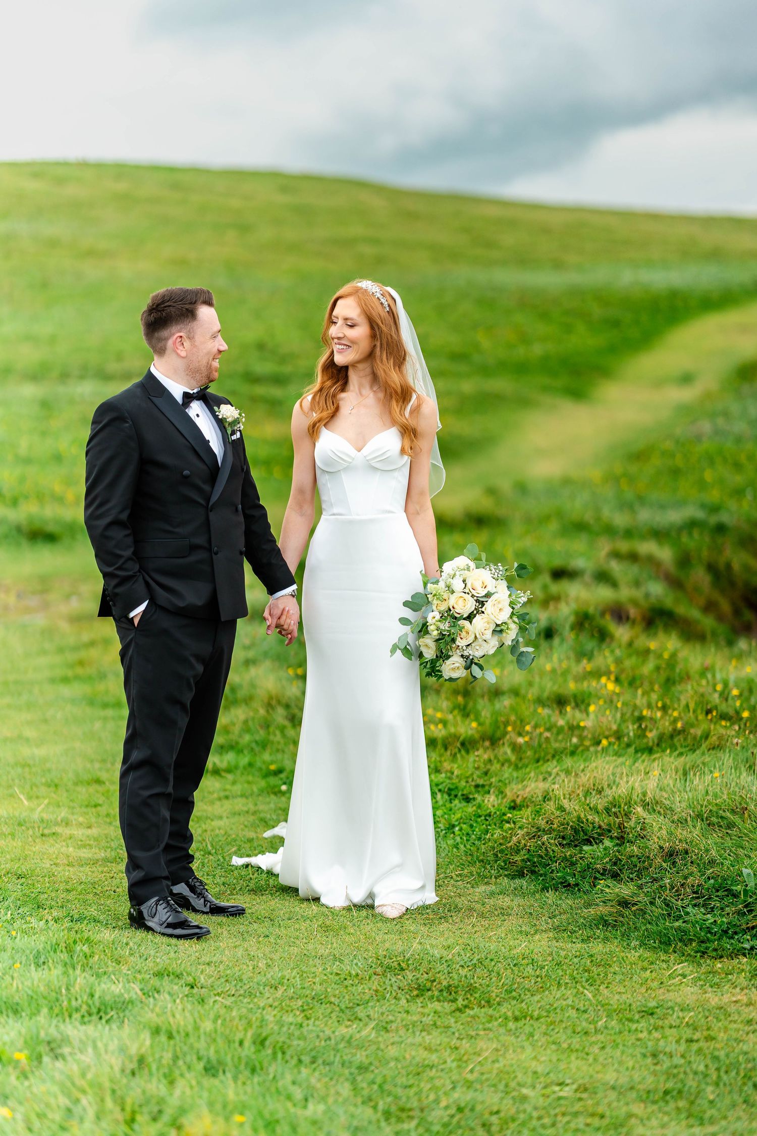 Wedding couple stands on rolling green hills during their coastal ceremony.