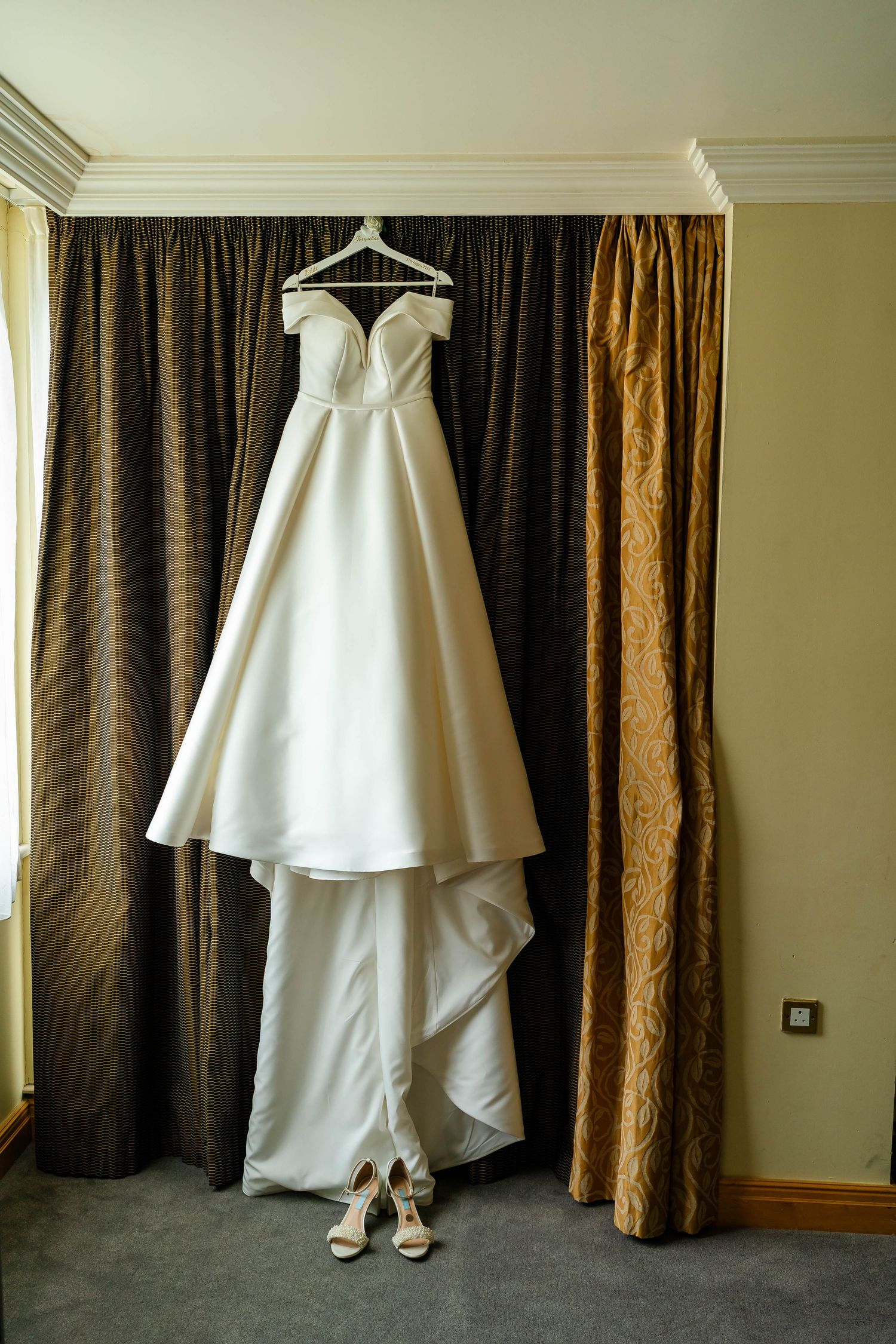 White wedding dress hanging on display against brown curtains in a hotel room.
