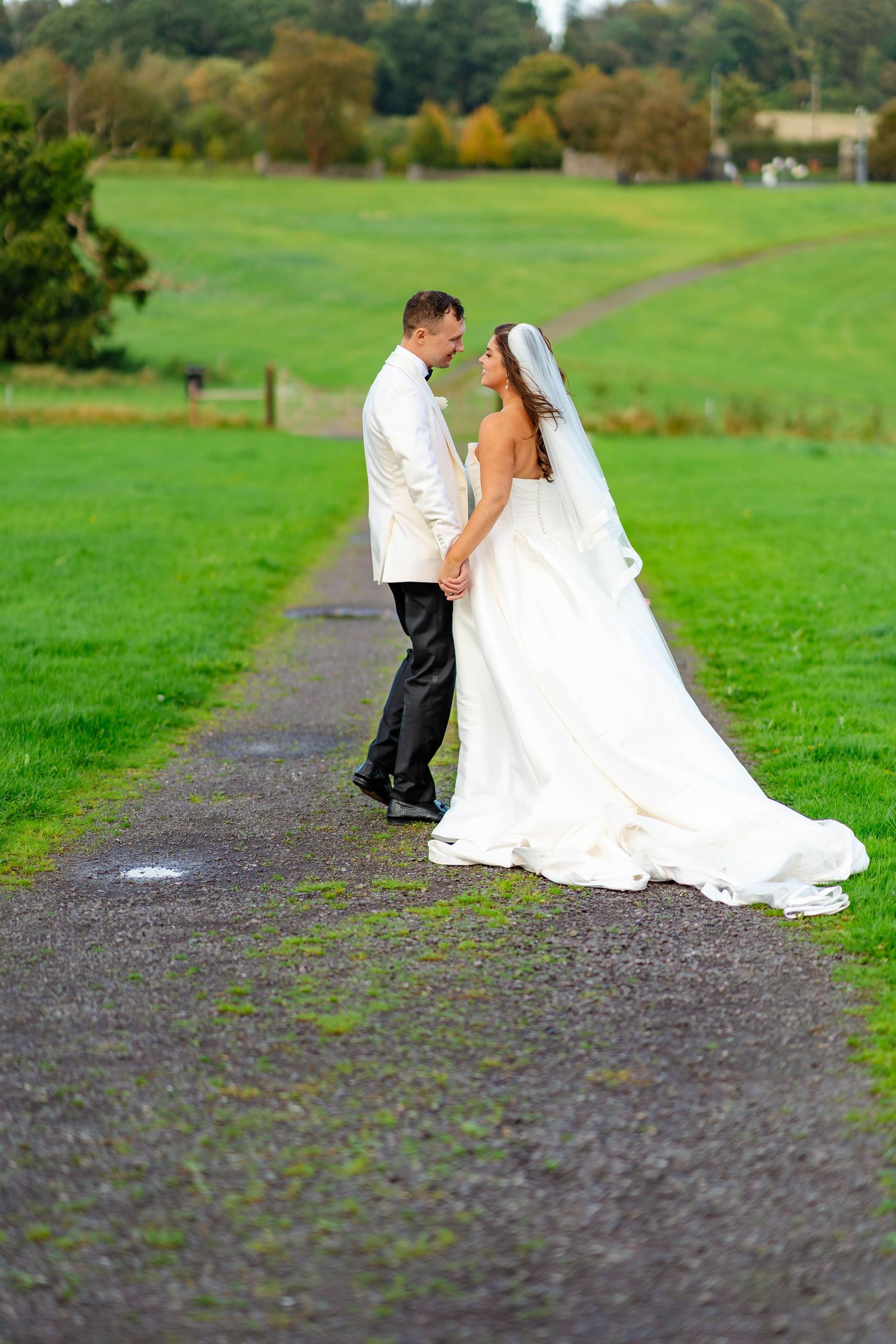 A couple in wedding attire stand together on a gravel path surrounded by lush green fields on their special day.