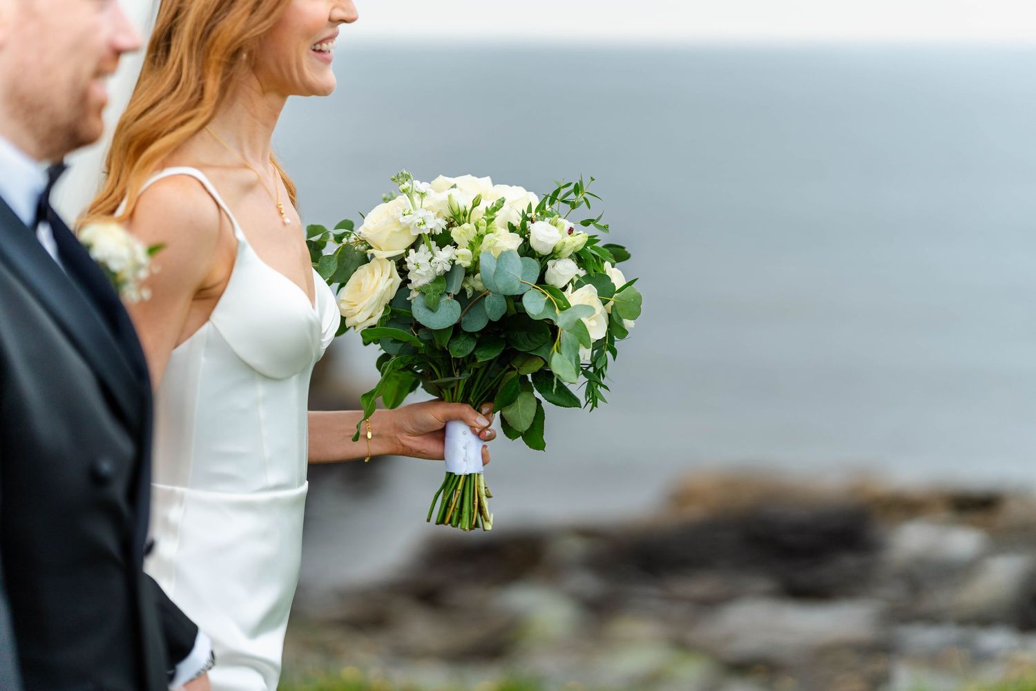 Elegant wedding bouquet of white flowers and greenery held against a coastal backdrop.