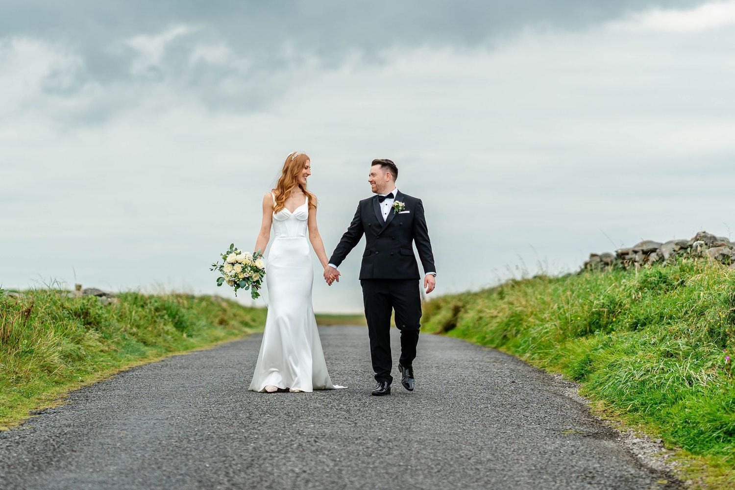 Newlyweds walk together on winding coastal road during overcast day.