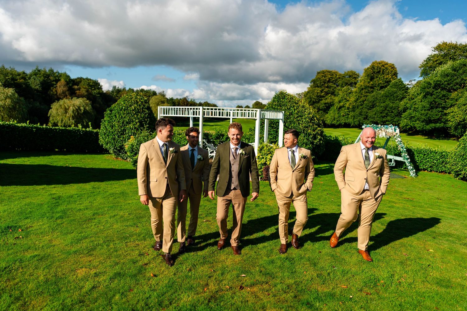 Wedding party in tan suits lines up for photos on rolling green lawn with white gazebo.