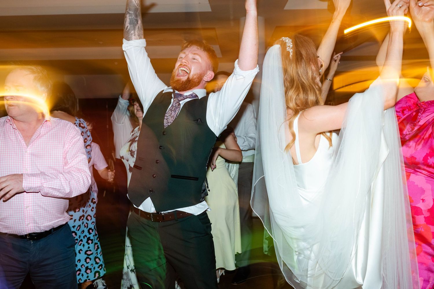 Wedding celebration with guests dancing with arms raised under warm lighting.