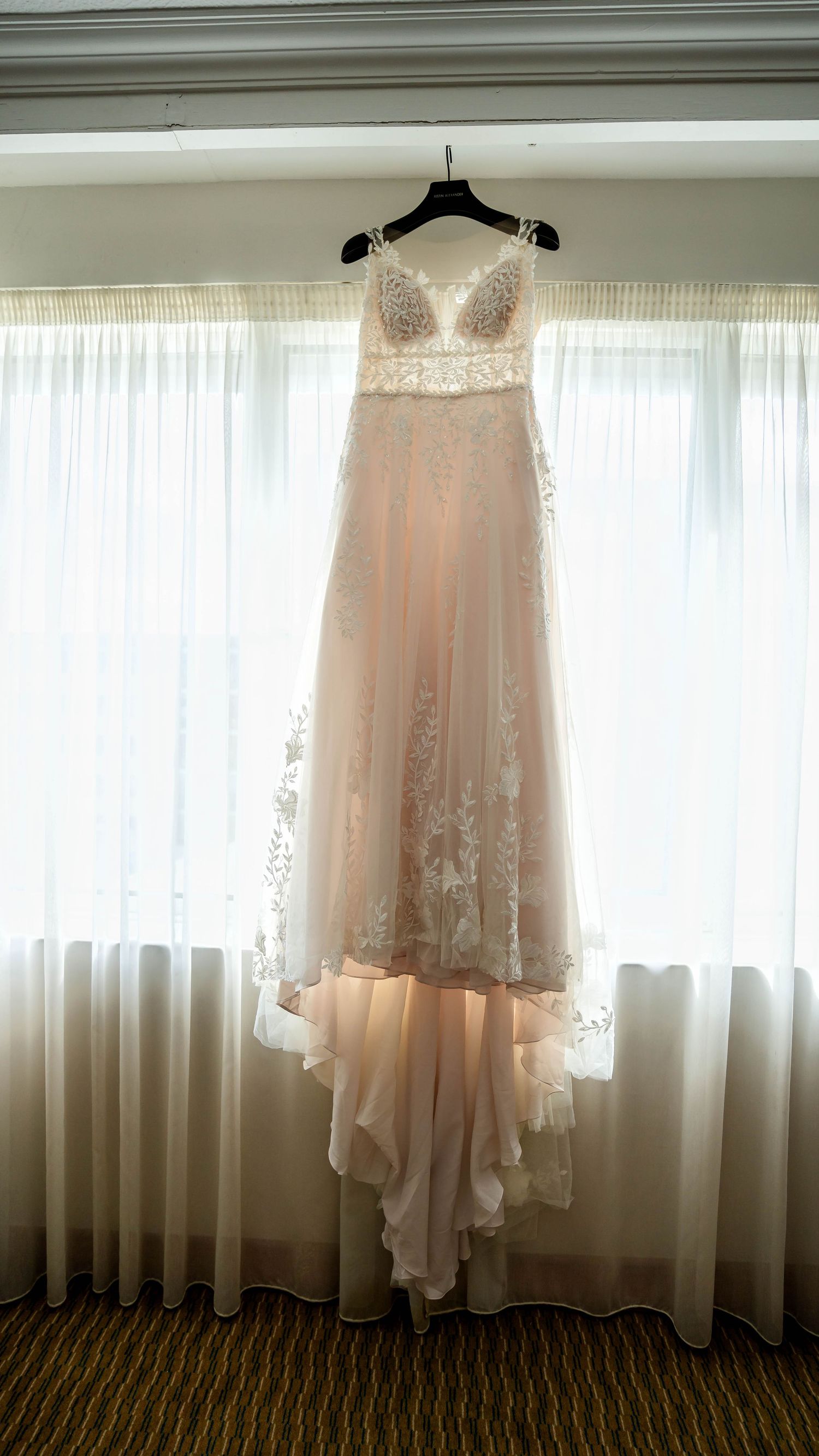 Delicate blush wedding dress hanging by window with soft natural lighting.