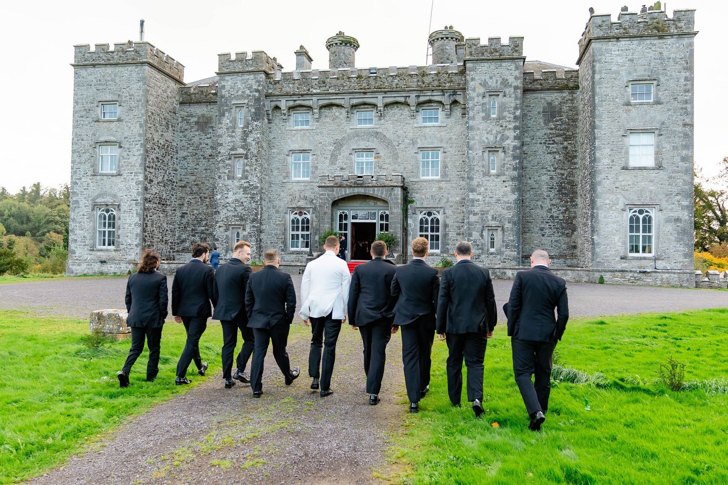 A group in formal attire walks toward a grand stone castle with turrets on a grassy lawn in Ireland.