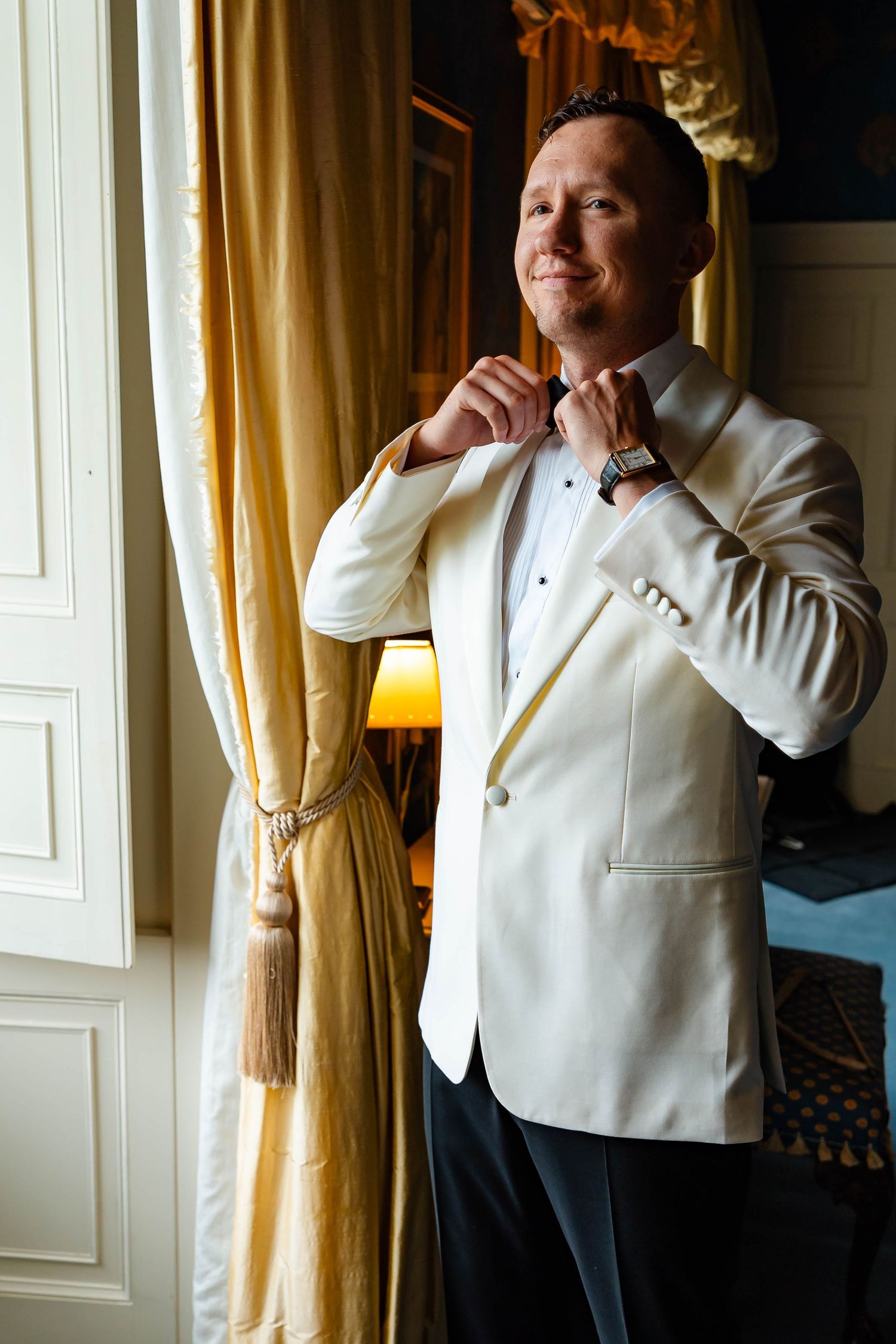 A groom in a white tuxedo jacket adjusts his bowtie while standing near elegant gold curtains in a formal setting.