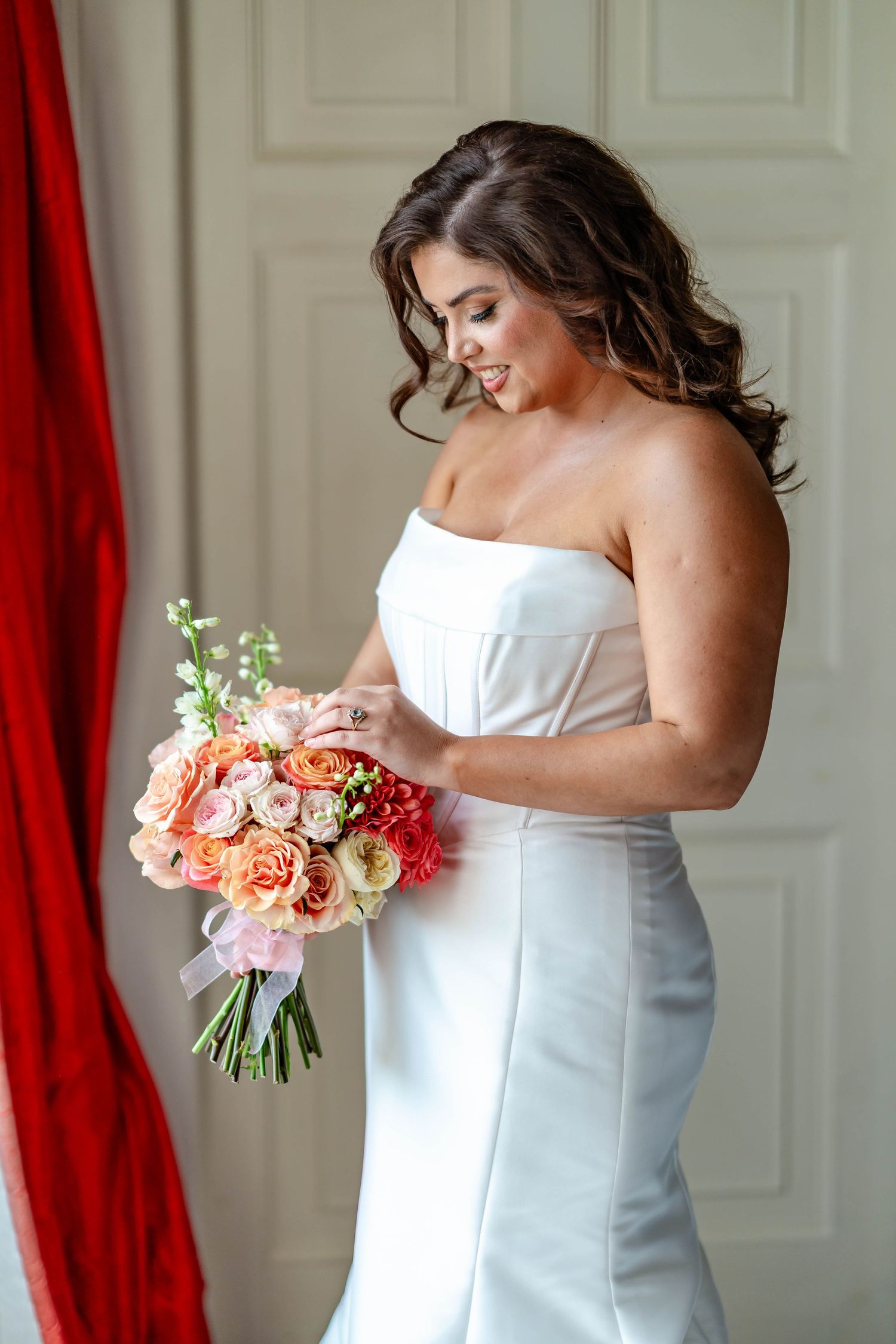 A bride in a white strapless gown holds a beautiful bouquet of coral and pink roses against a red curtain backdrop.