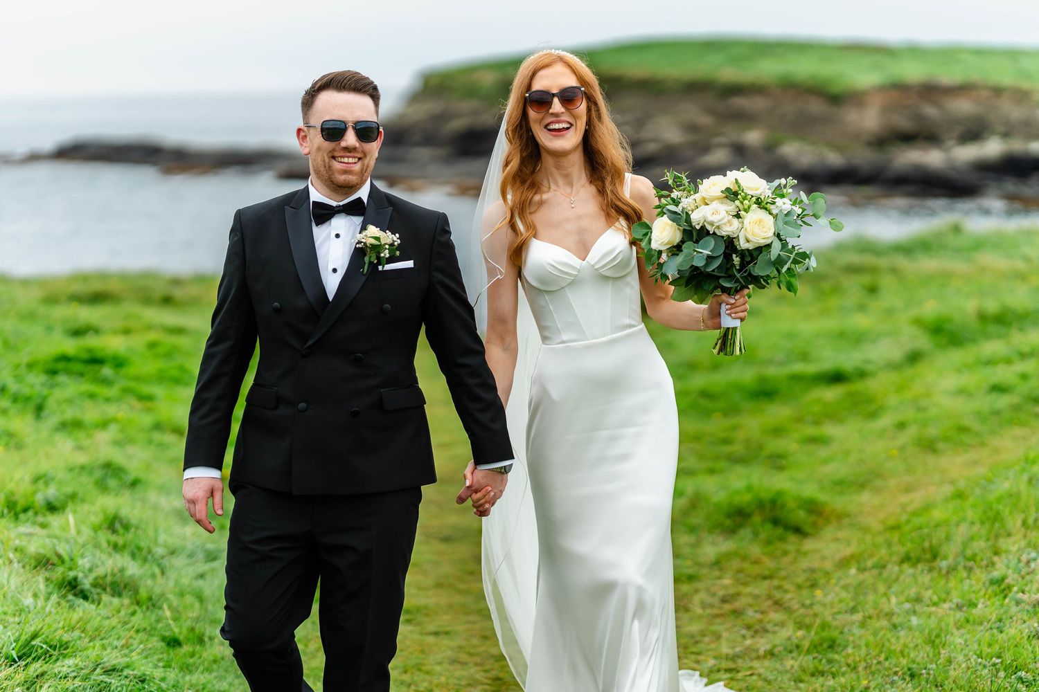 Newlyweds walk together on lush green coastal cliffs while holding hands and bouquet.