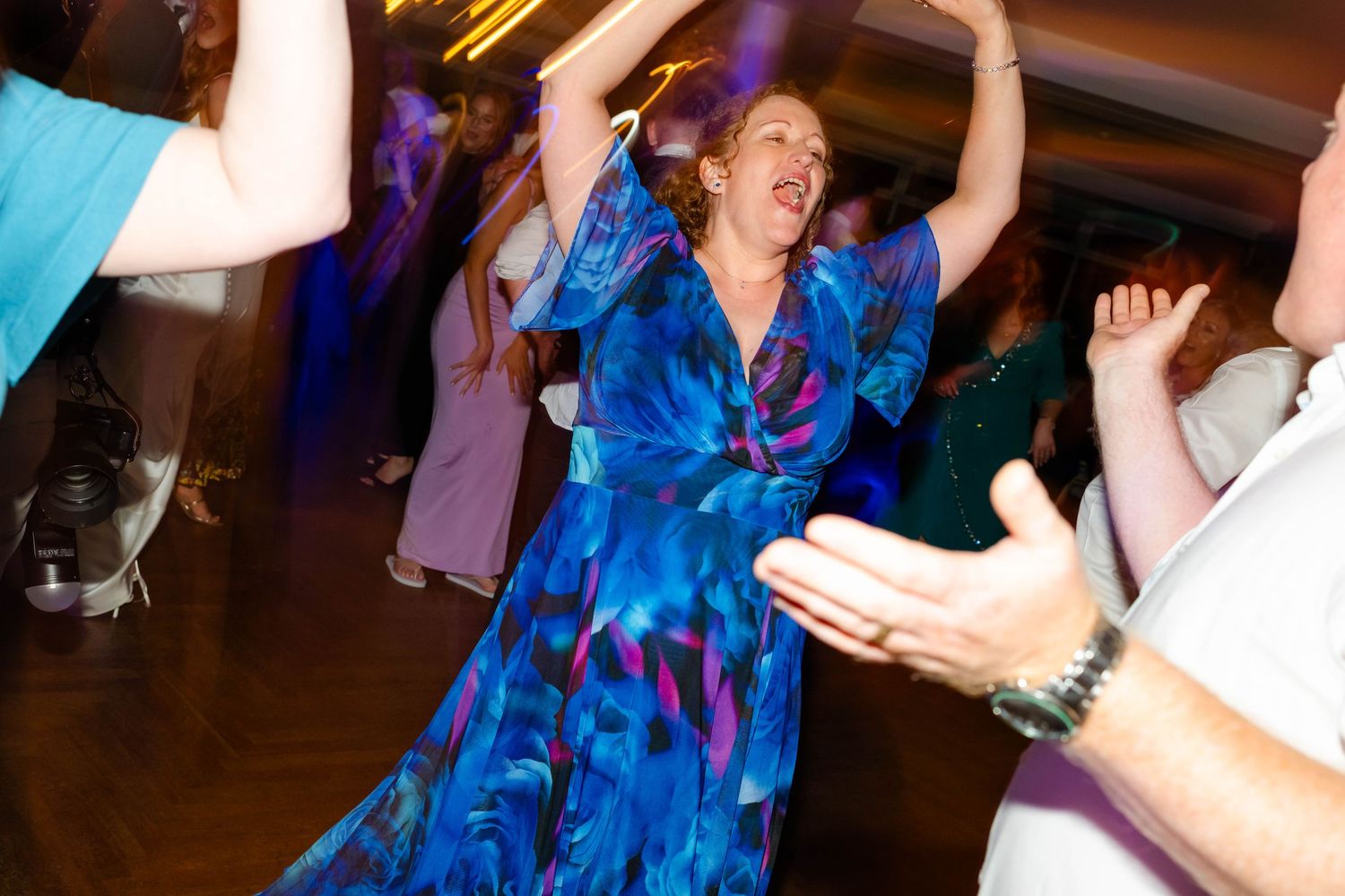Woman in vibrant blue patterned dress dances enthusiastically at wedding reception.