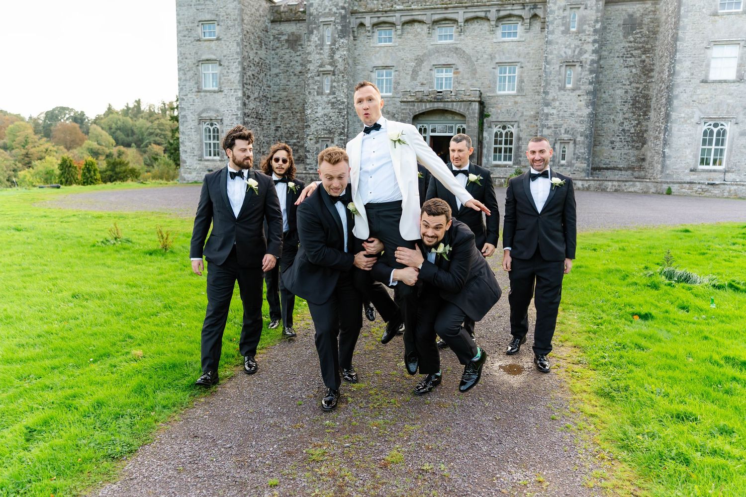 Wedding party members in black tuxedos walk and pose together outside a grand stone castle on a green lawn.