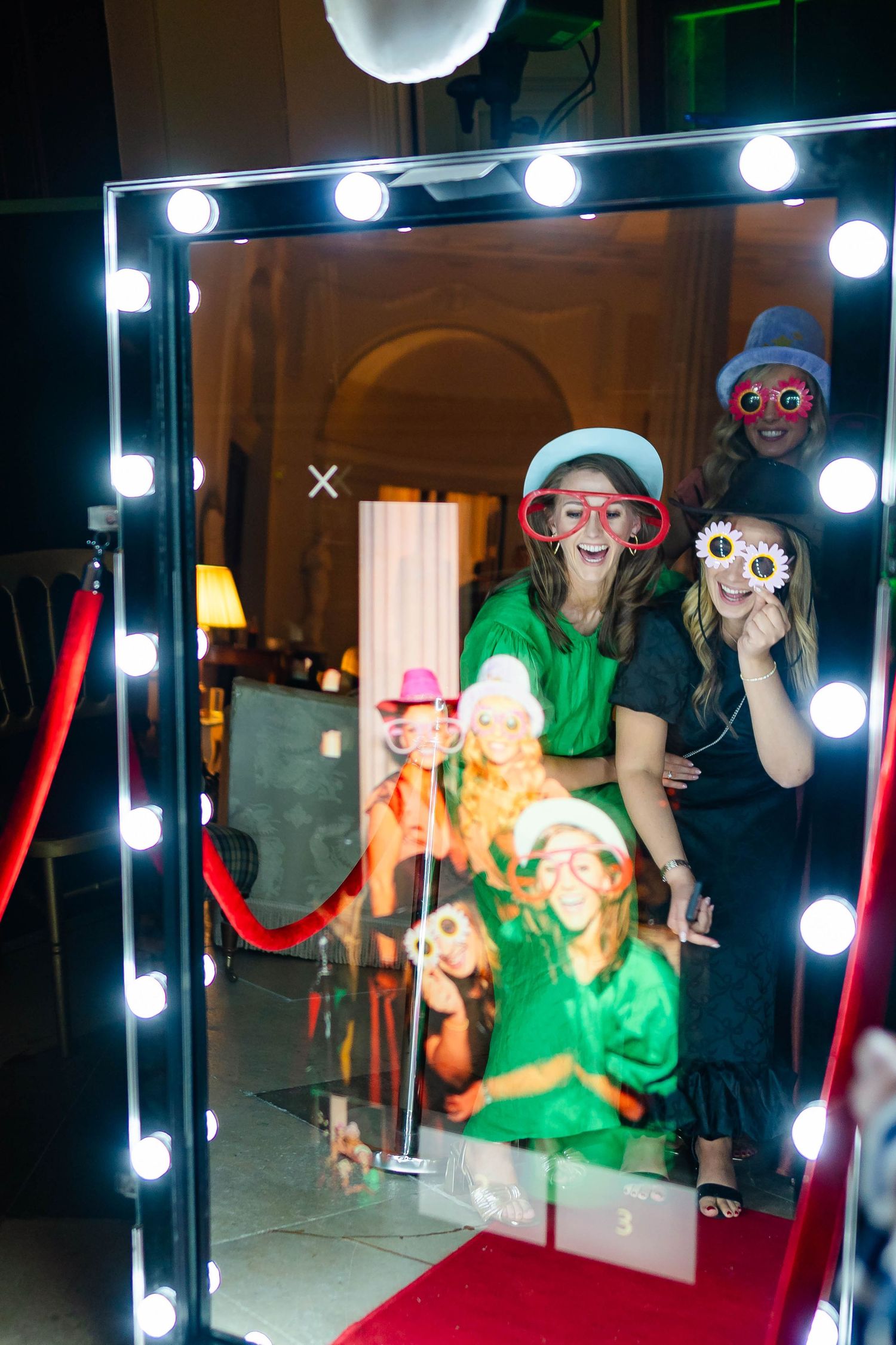 People in colorful costumes pose playfully in a mirror reflection with bright vanity lights and red fabric visible.