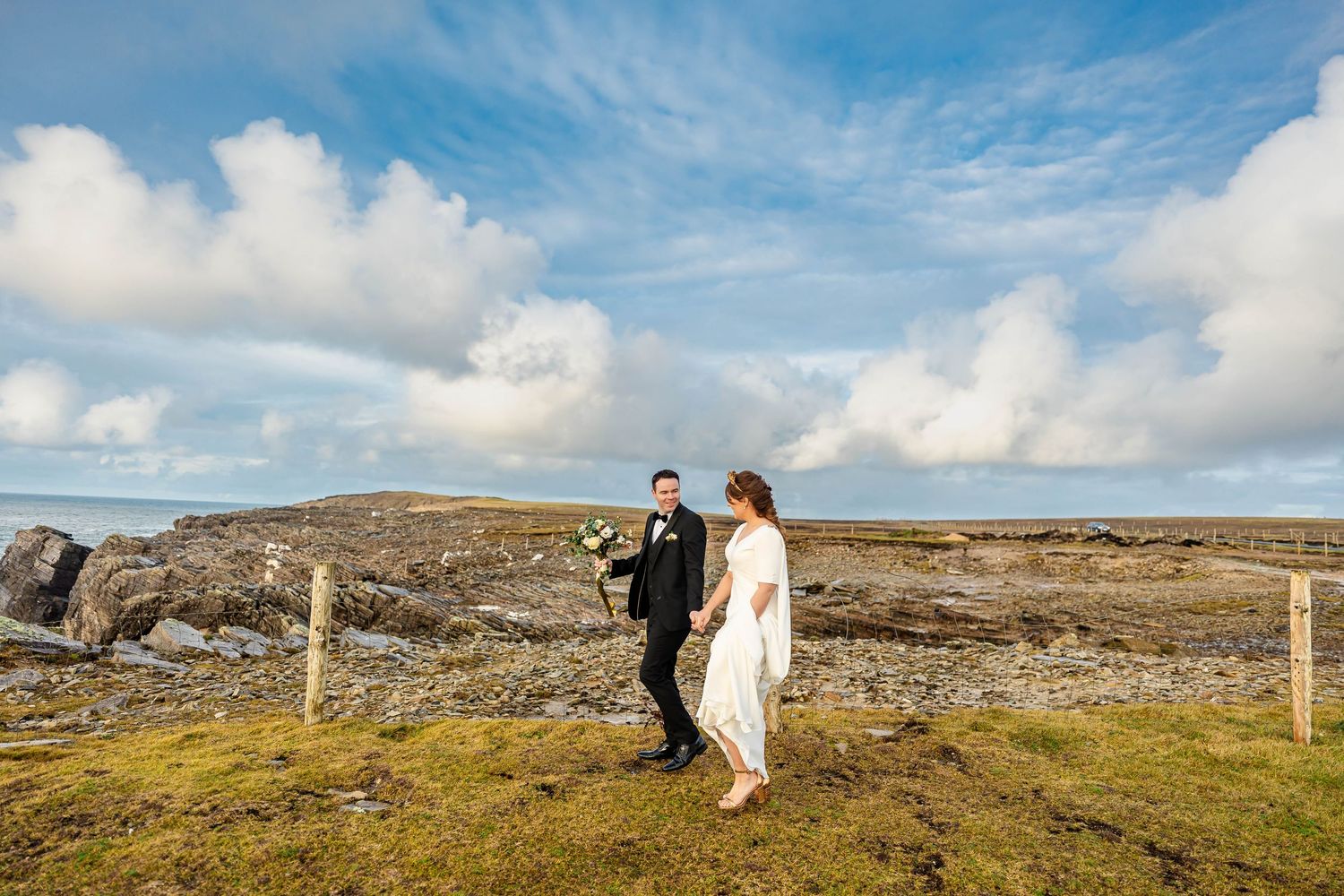 Couple strolls hand in hand across windswept moorland with fence posts and blue sky.