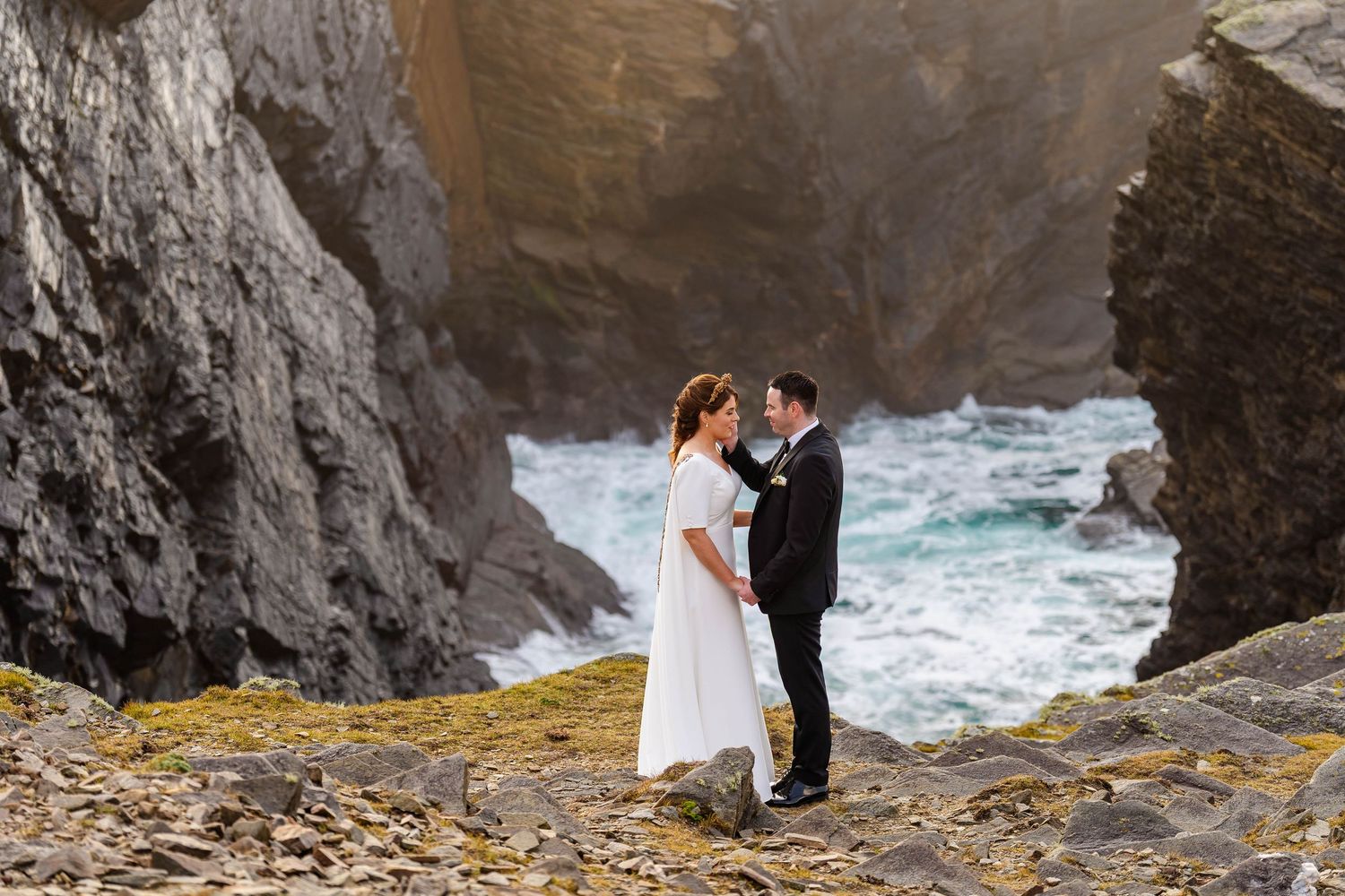 Wedding couple embraces on rocky coastline with dramatic ocean waves and cliffs.