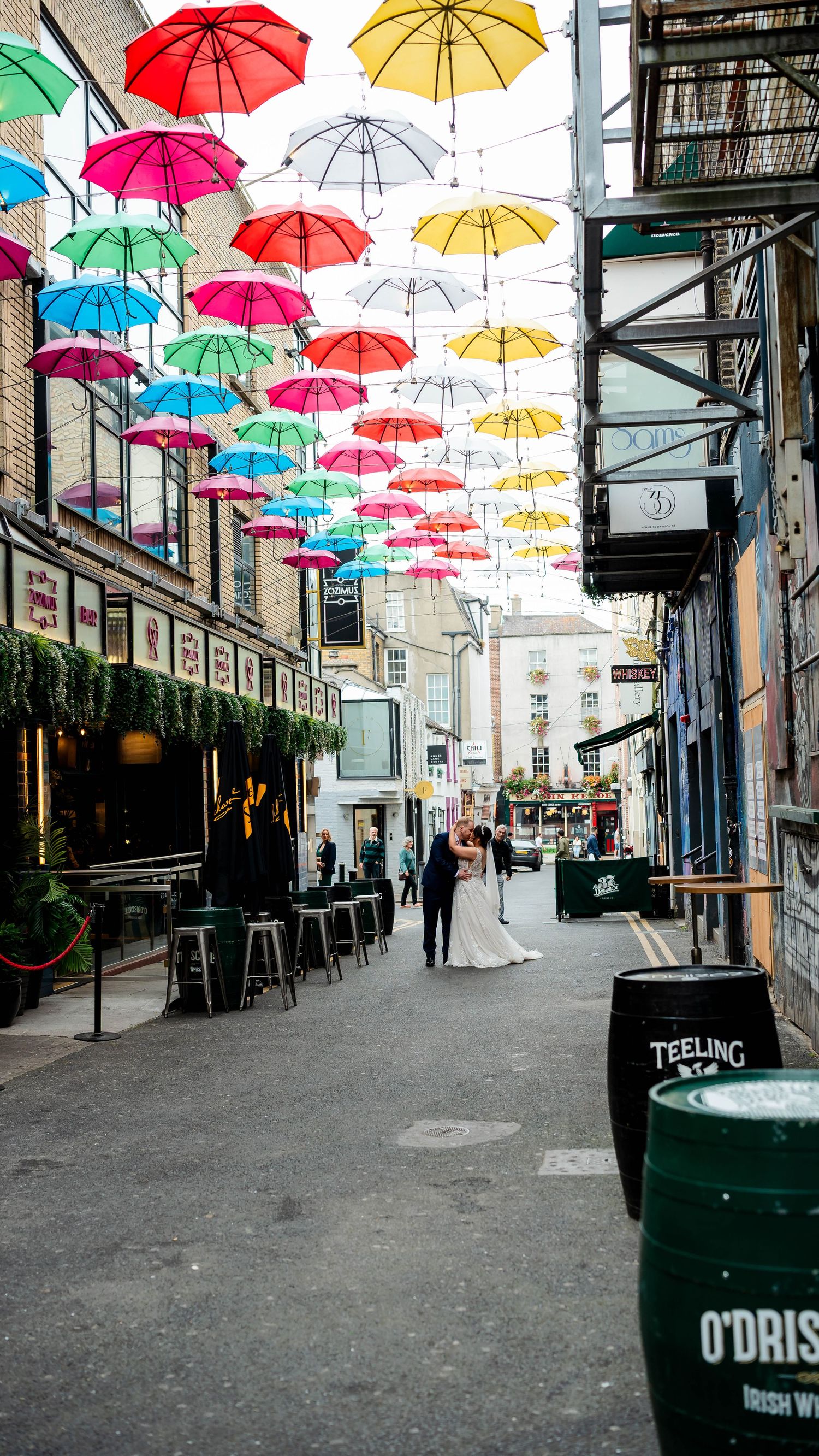 Colorful umbrellas hang above a charming cobblestone street lined with shops and restaurants.