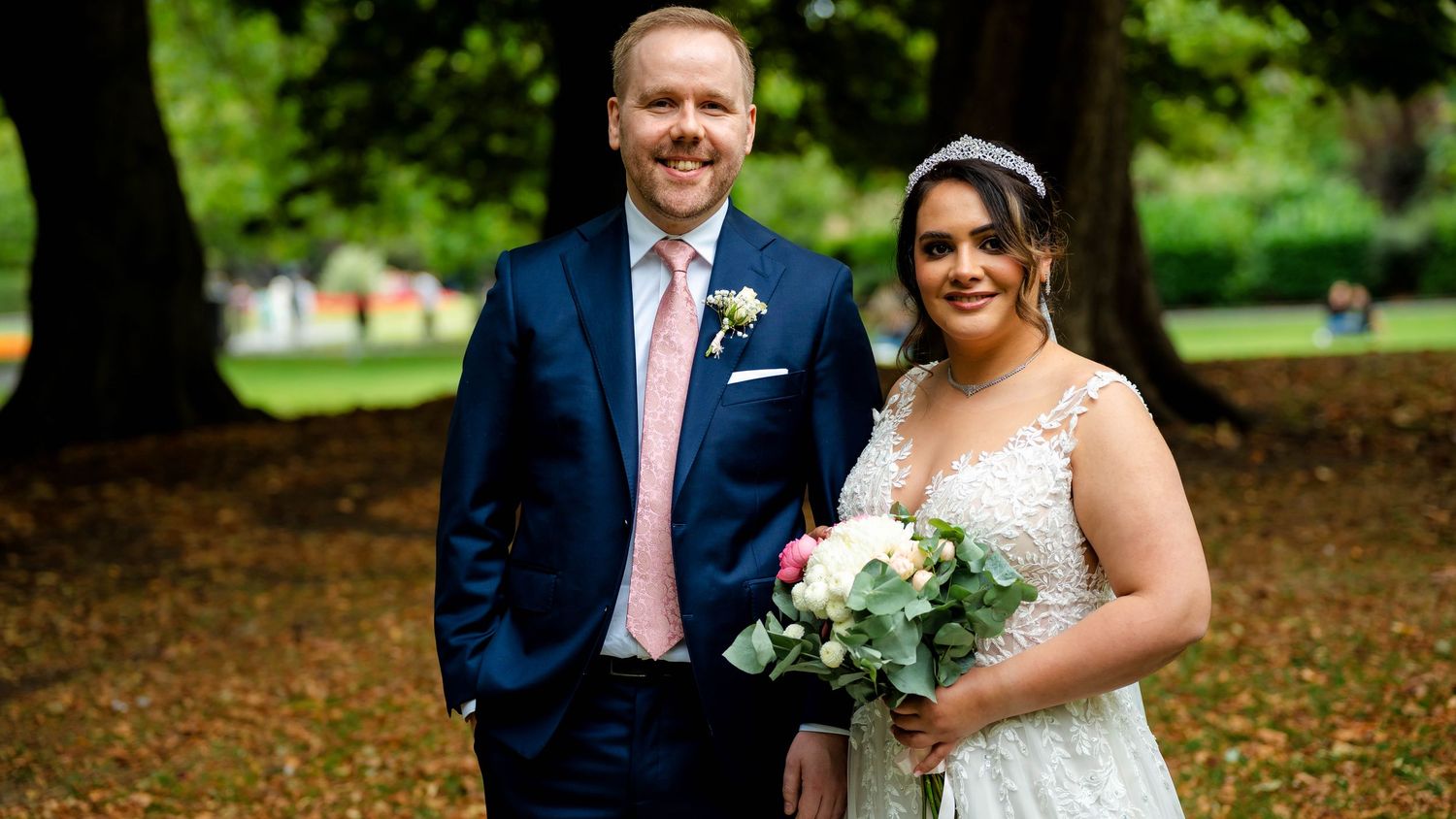A wedding party stands together in a scenic park setting during fall.