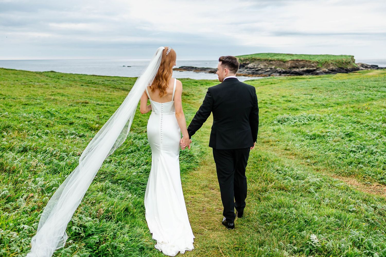 Newlyweds walk hand in hand across a dramatic coastal clifftop with flowing veil in the wind.
