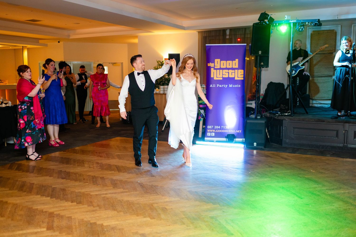 Newlyweds dancing together on parquet floor with colorful lighting and band setup visible.