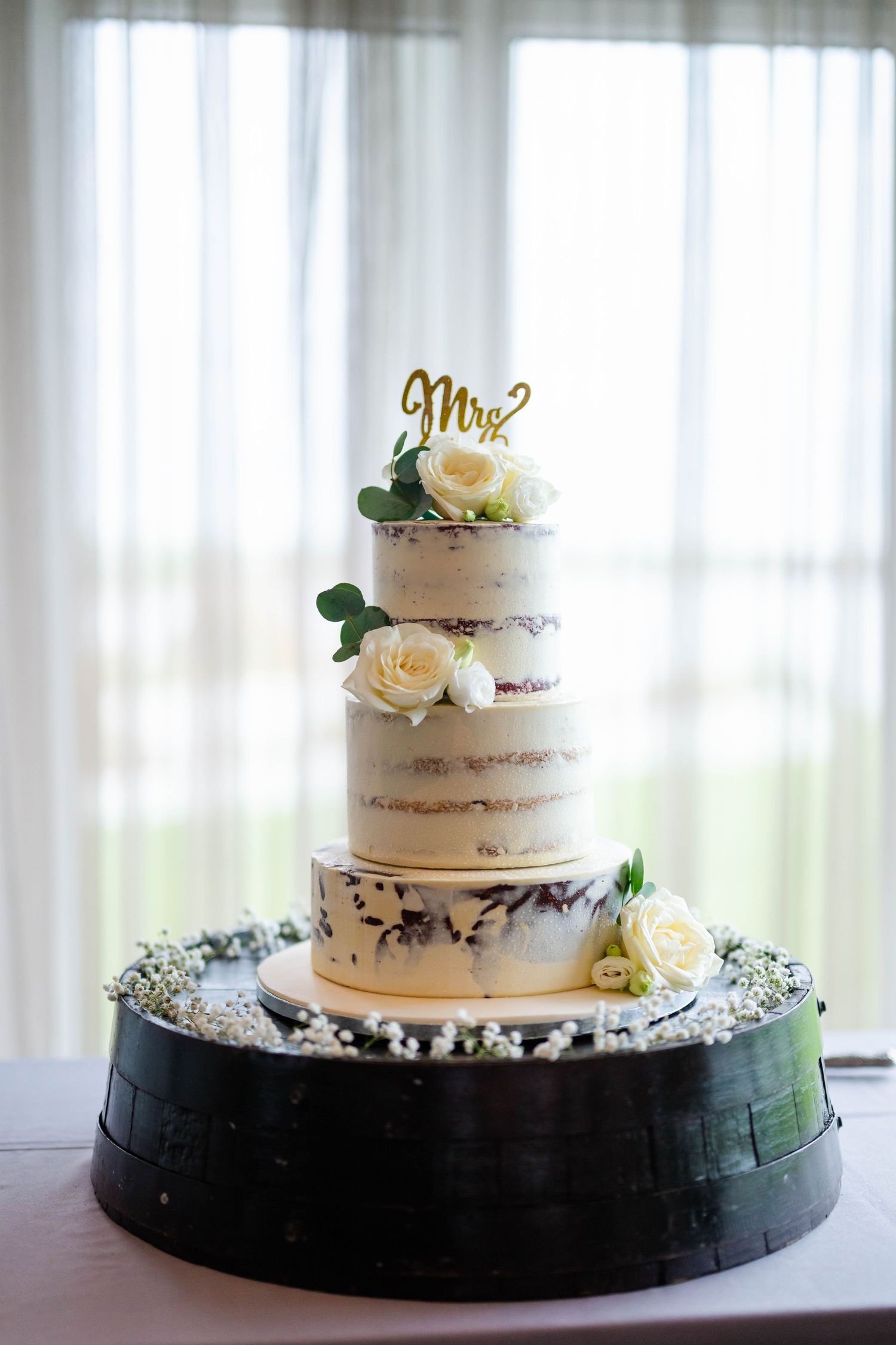 Elegant three-tier white wedding cake with floral decorations displayed against white curtains.