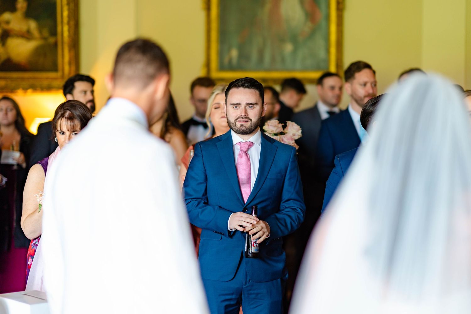 Wedding guests in blue suits and formal attire gather at an elegant indoor reception venue.
