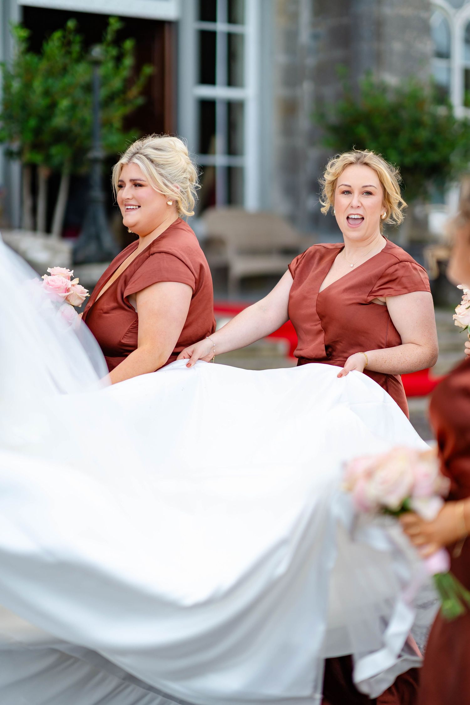 Bridesmaids in burgundy dresses help fluff and arrange a white wedding dress train in an outdoor setting.