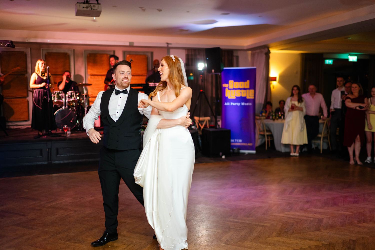Couple sharing their first dance on wooden floor at wedding reception with band in background.