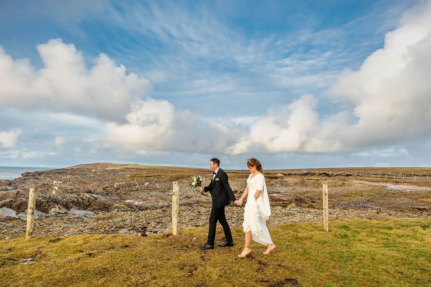 Couple walking along fence line on rugged coastal landscape under blue skies.