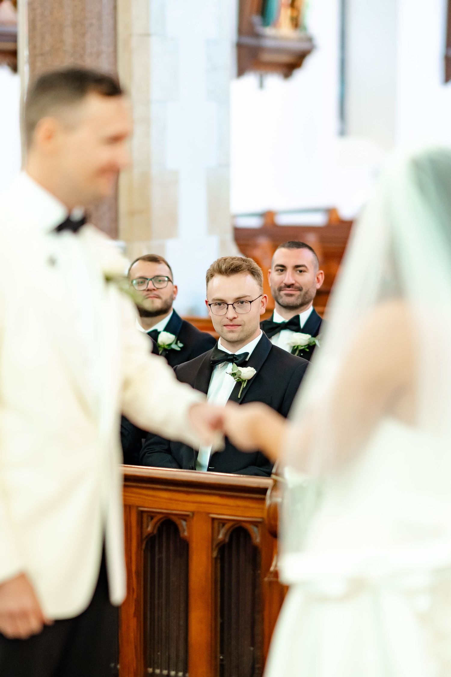 Wedding guests in formal attire watch the ceremony from wooden church pews.