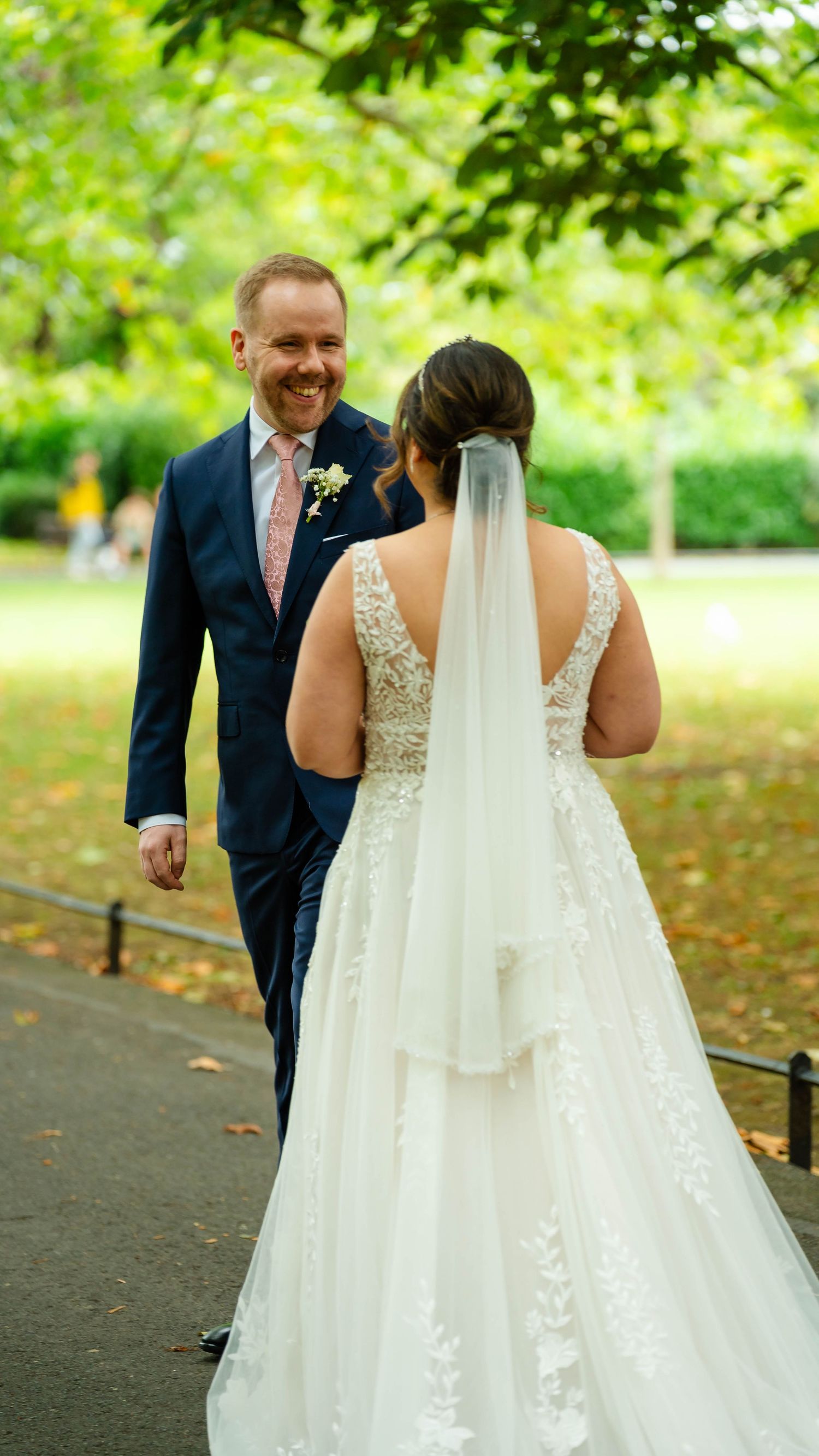 First look wedding moment captured on a tree-lined path with bride in lace gown and groom in navy suit.