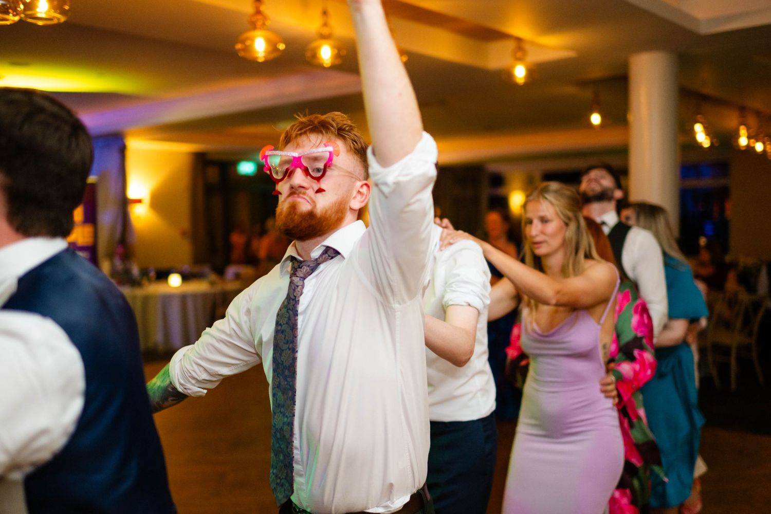 Wedding guests dancing in formation under warm ambient lighting at an indoor reception.