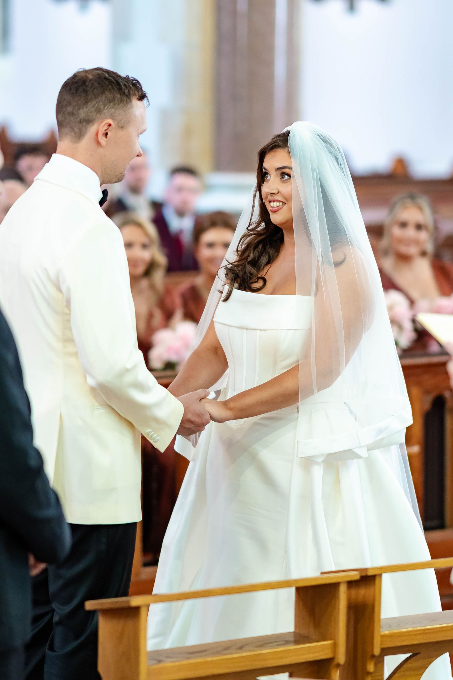 A couple exchanges vows during their wedding ceremony in a church with guests watching in the background.