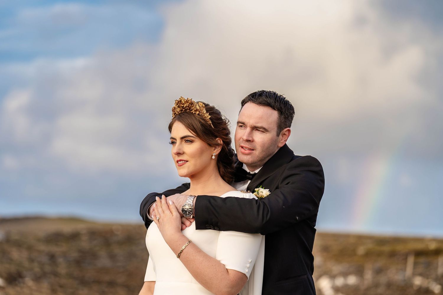 Wedding couple embracing against dramatic cloudy sky with rainbow in background.