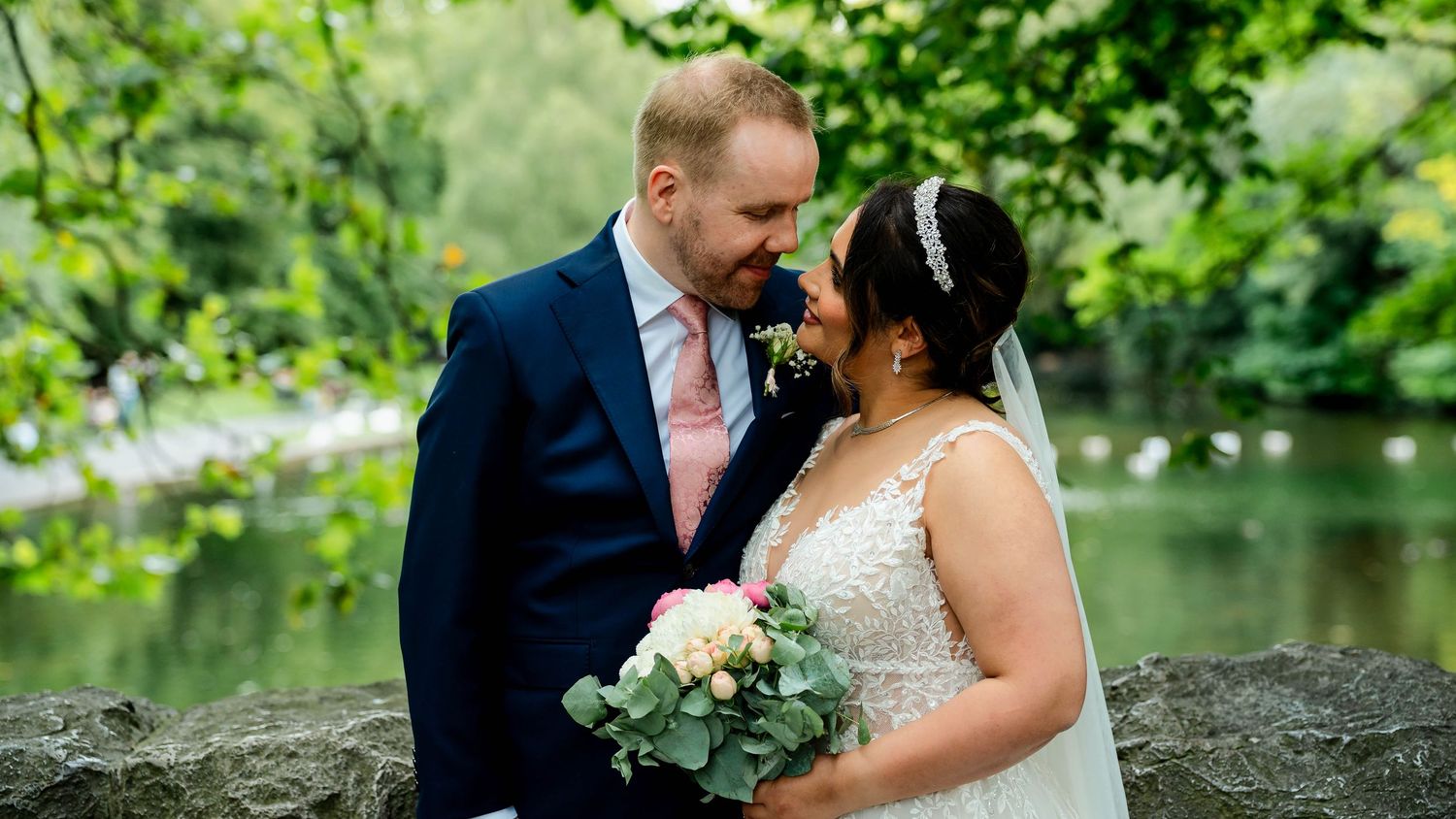 A couple shares an intimate moment by a pond in a lush garden setting.