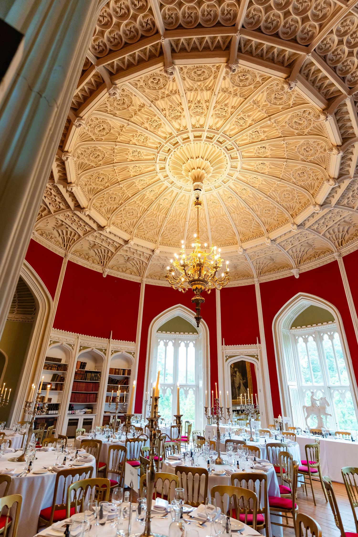 Ornate white ceiling with intricate plasterwork and chandelier in an elegant red dining room with Gothic windows.