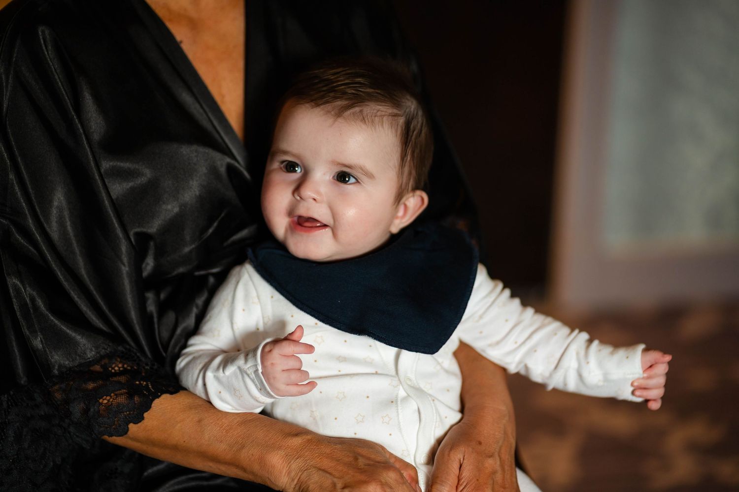 Baby in white clothing reaching out while being held against a warm-toned background.