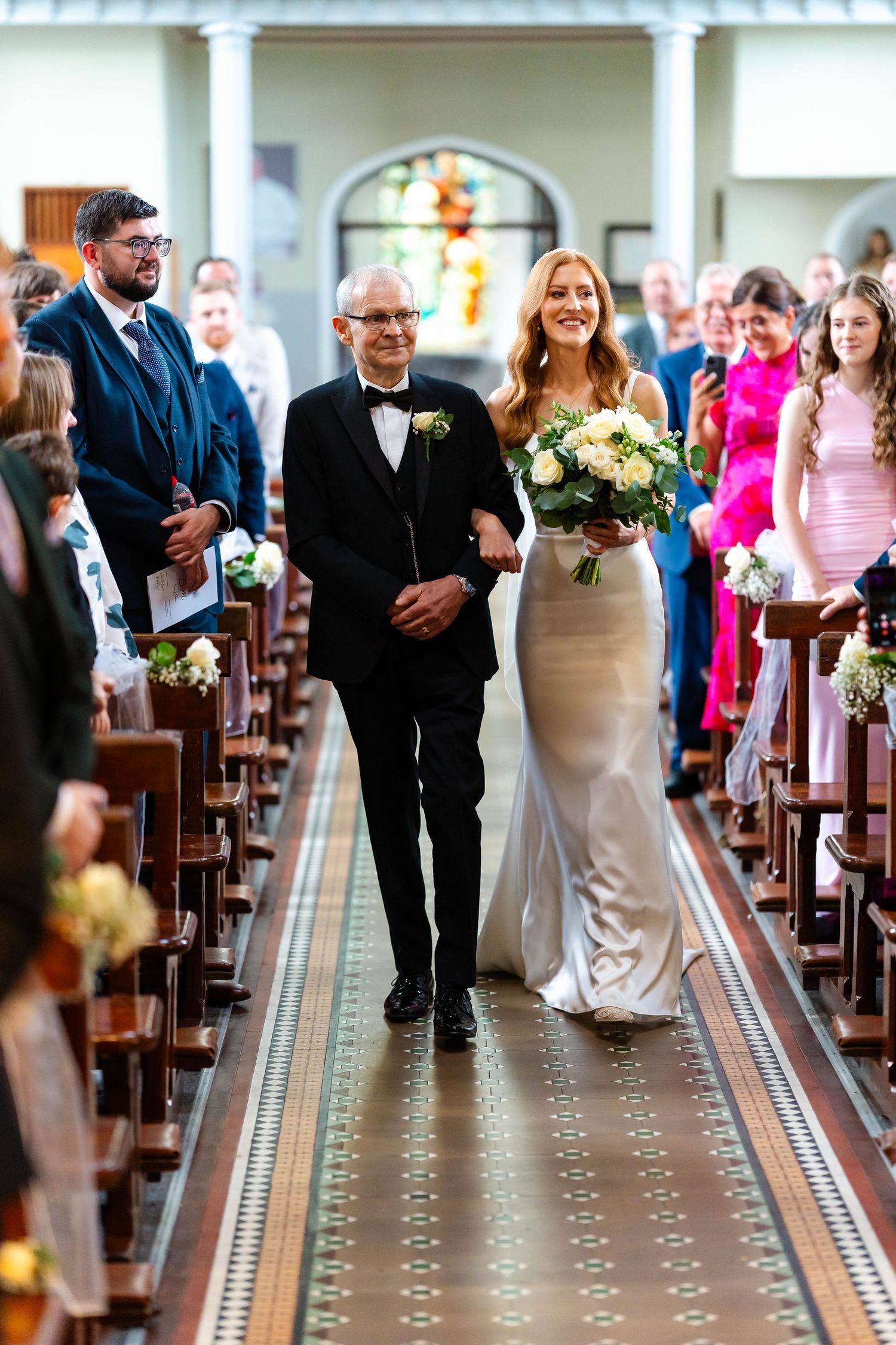 Wedding procession walking down ornate church aisle with decorative floor pattern and wooden pews.