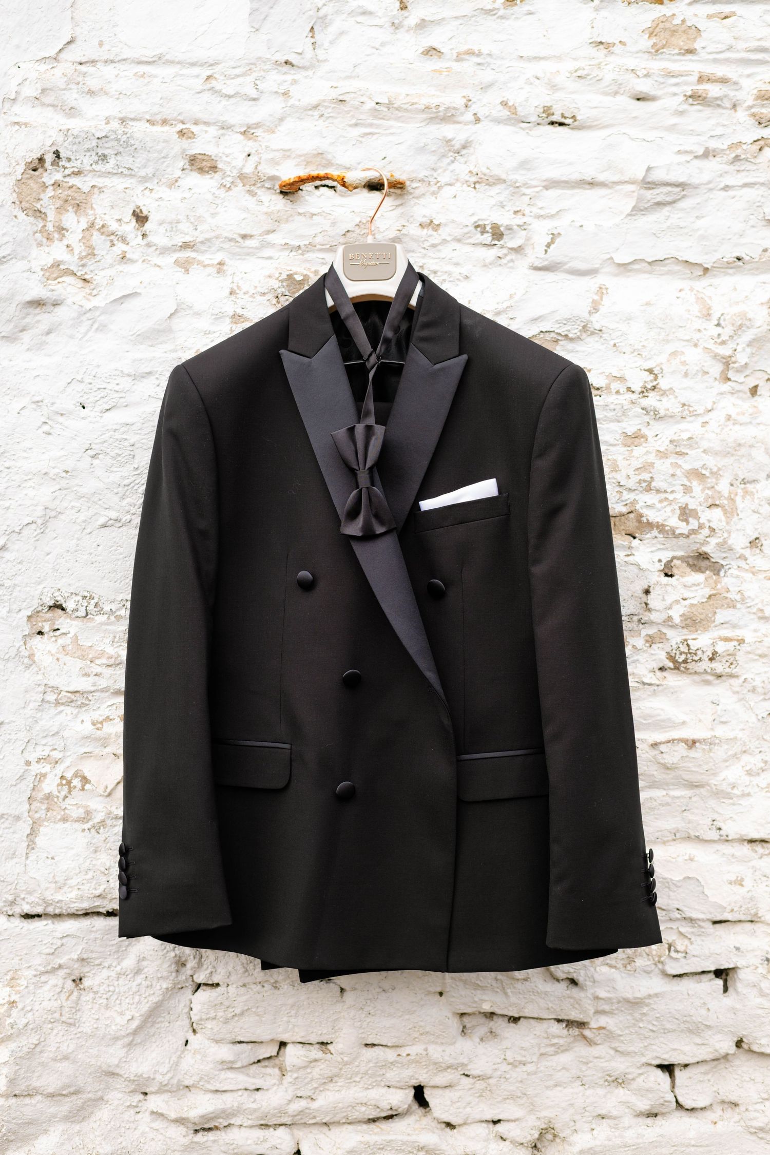 Classic black tuxedo jackets hanging against a textured white brick wall.