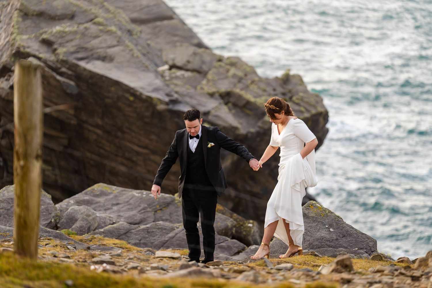 Wedding couple walking along dramatic coastal cliffs with ocean waves below.