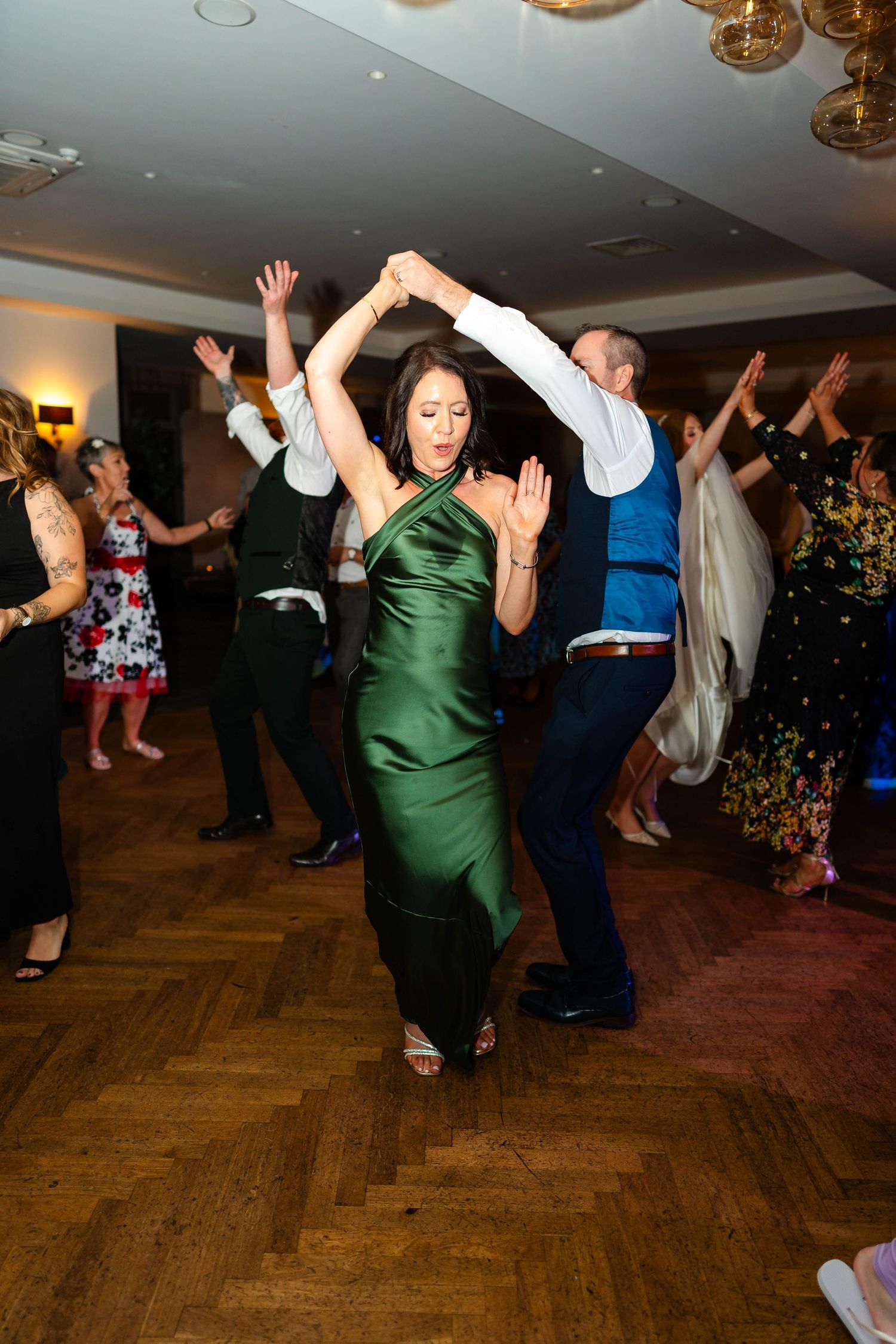 Guests dancing at wedding reception with woman in emerald green dress being twirled on parquet floor.