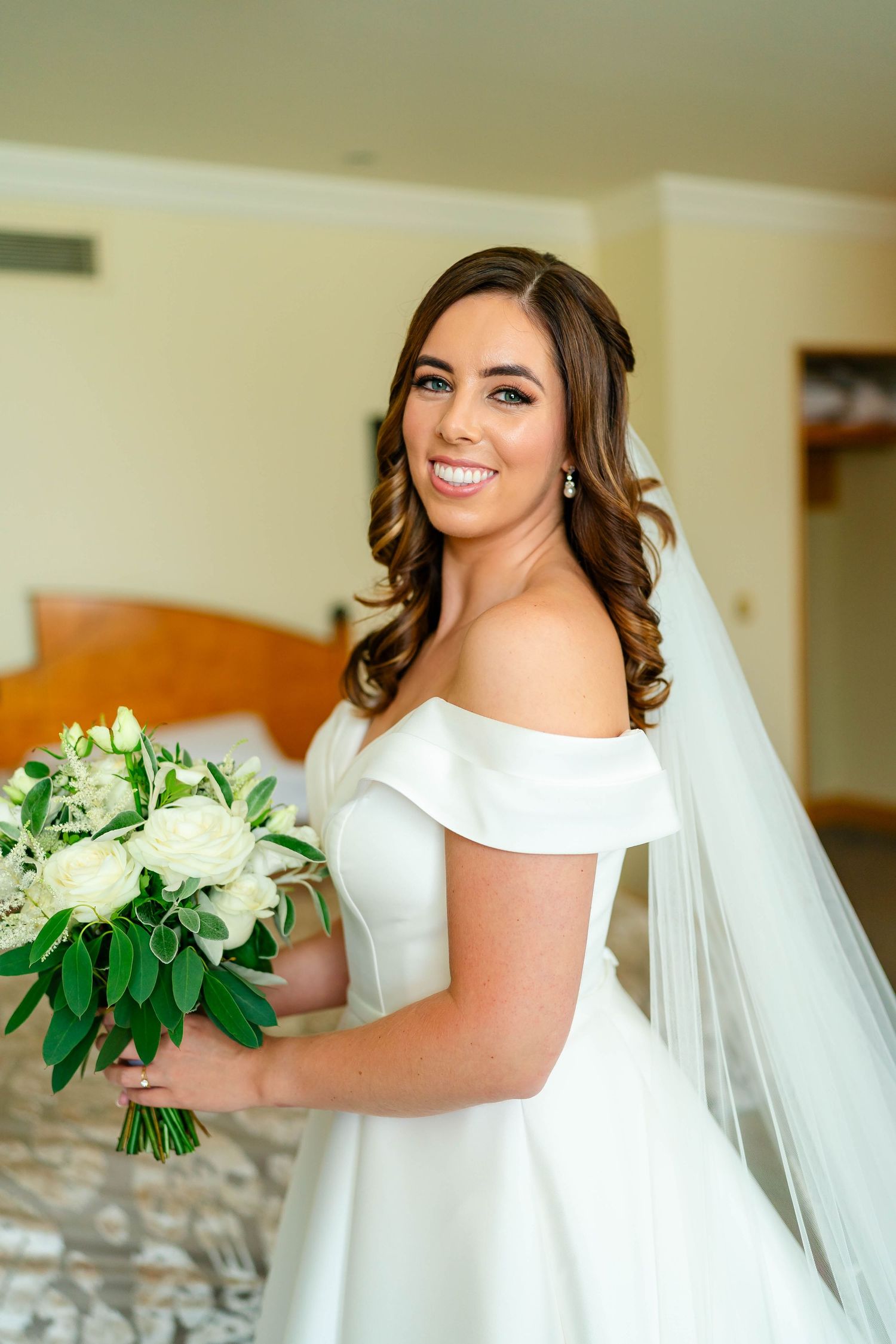A bride in an elegant off-shoulder white dress holds a bouquet of white flowers and greenery.