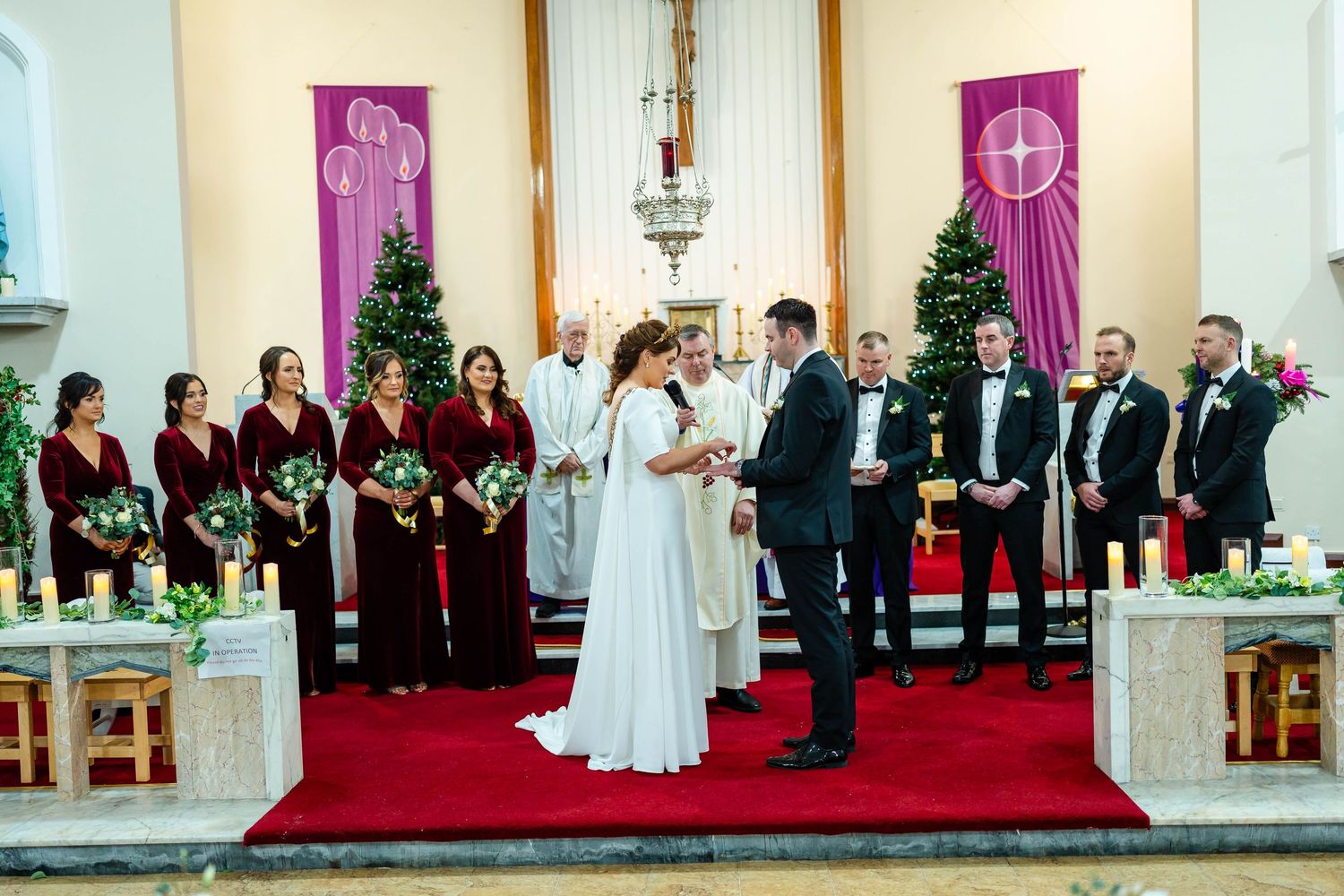 Wedding ceremony in progress at a church with purple decor and Christmas trees in the background.