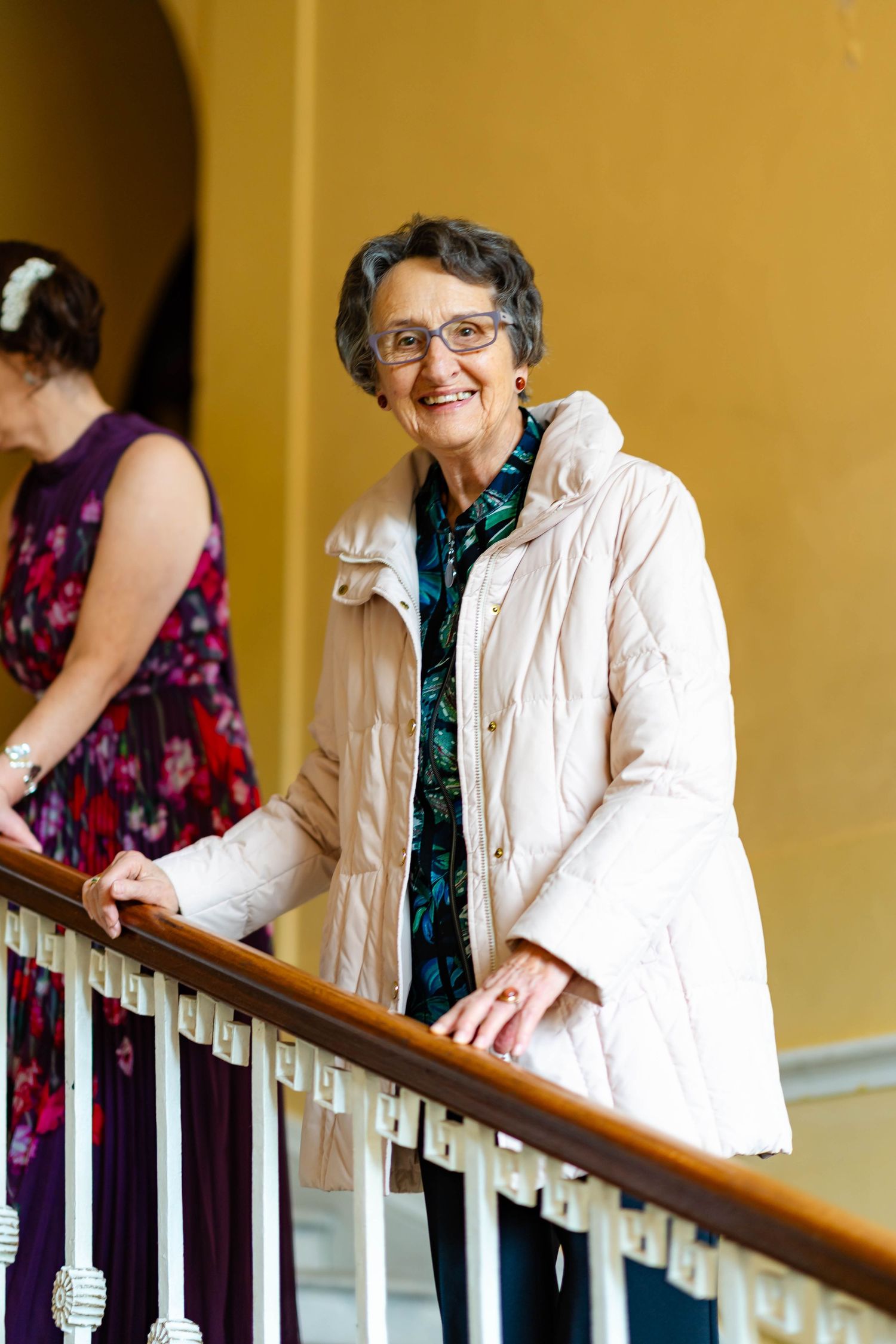 A person in a cream-colored winter coat and glasses stands smiling on a staircase with a wooden banister.